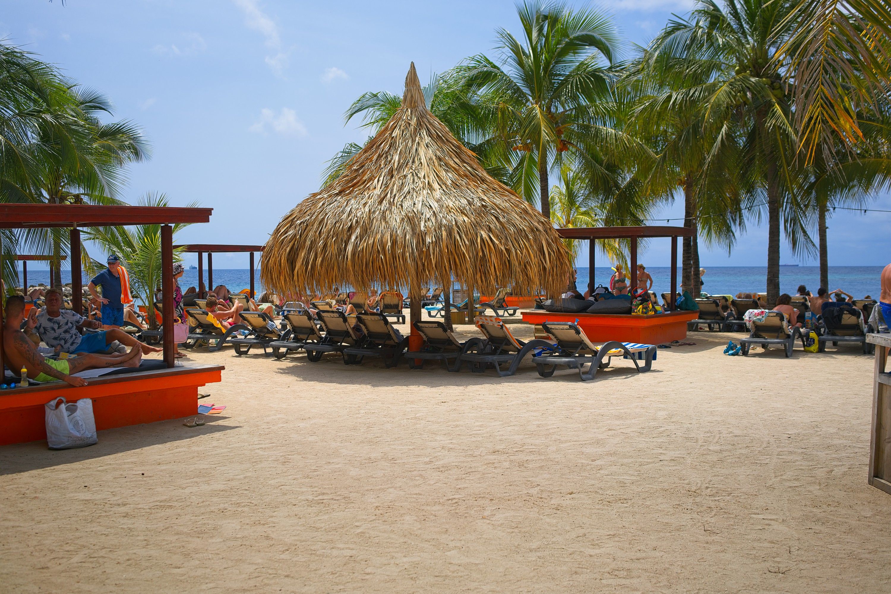 Busy beach scene at Jan Thiel Beach, Curacao, featuring vacationers relaxing under a large thatched umbrella and palm trees, with ocean views in the background.