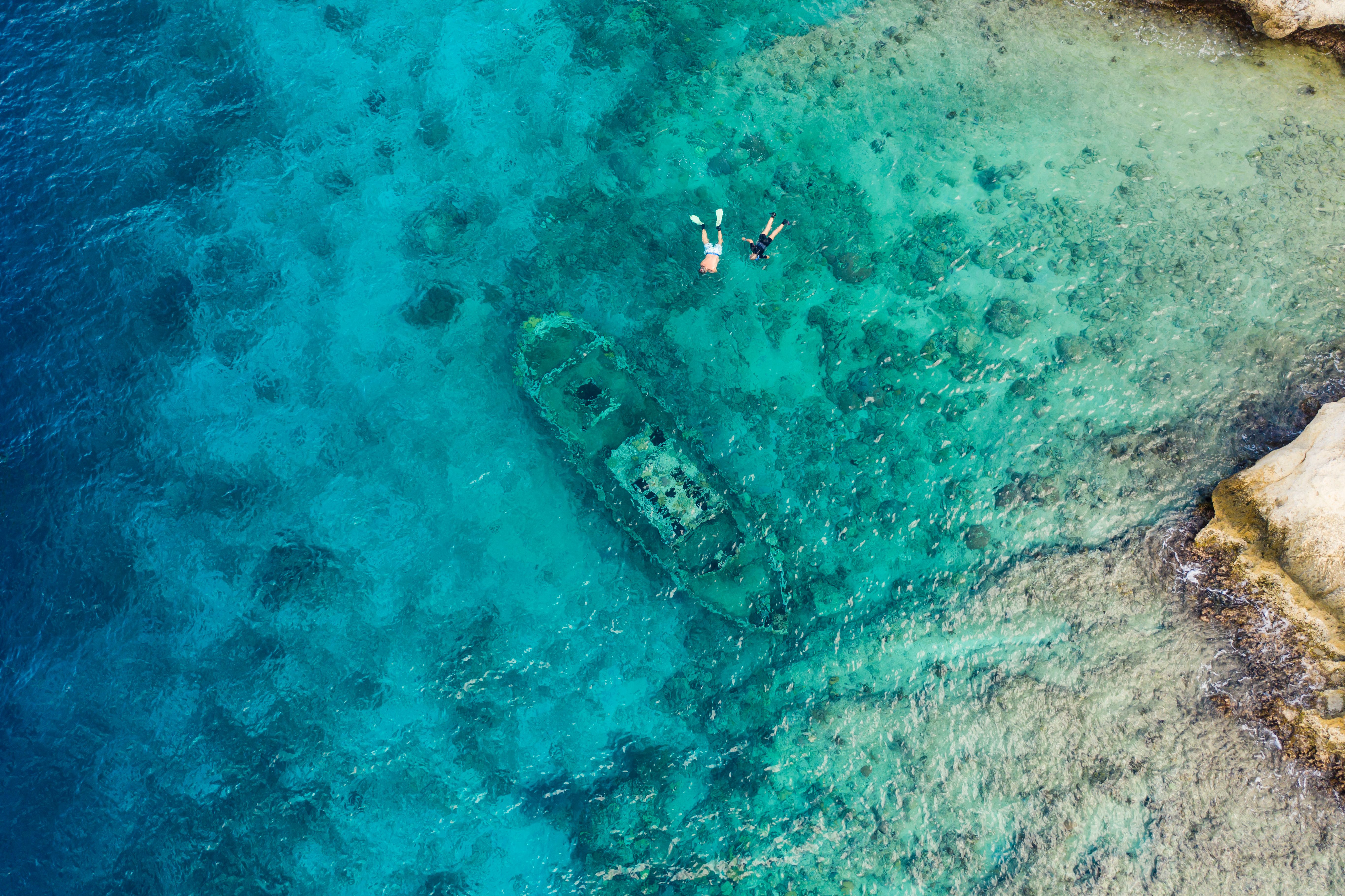 Aerial view of snorkelers exploring a sunken tugboat in the clear turquoise waters off Tugboat Beach, Curacao, with visible coral reefs and rocky shore.