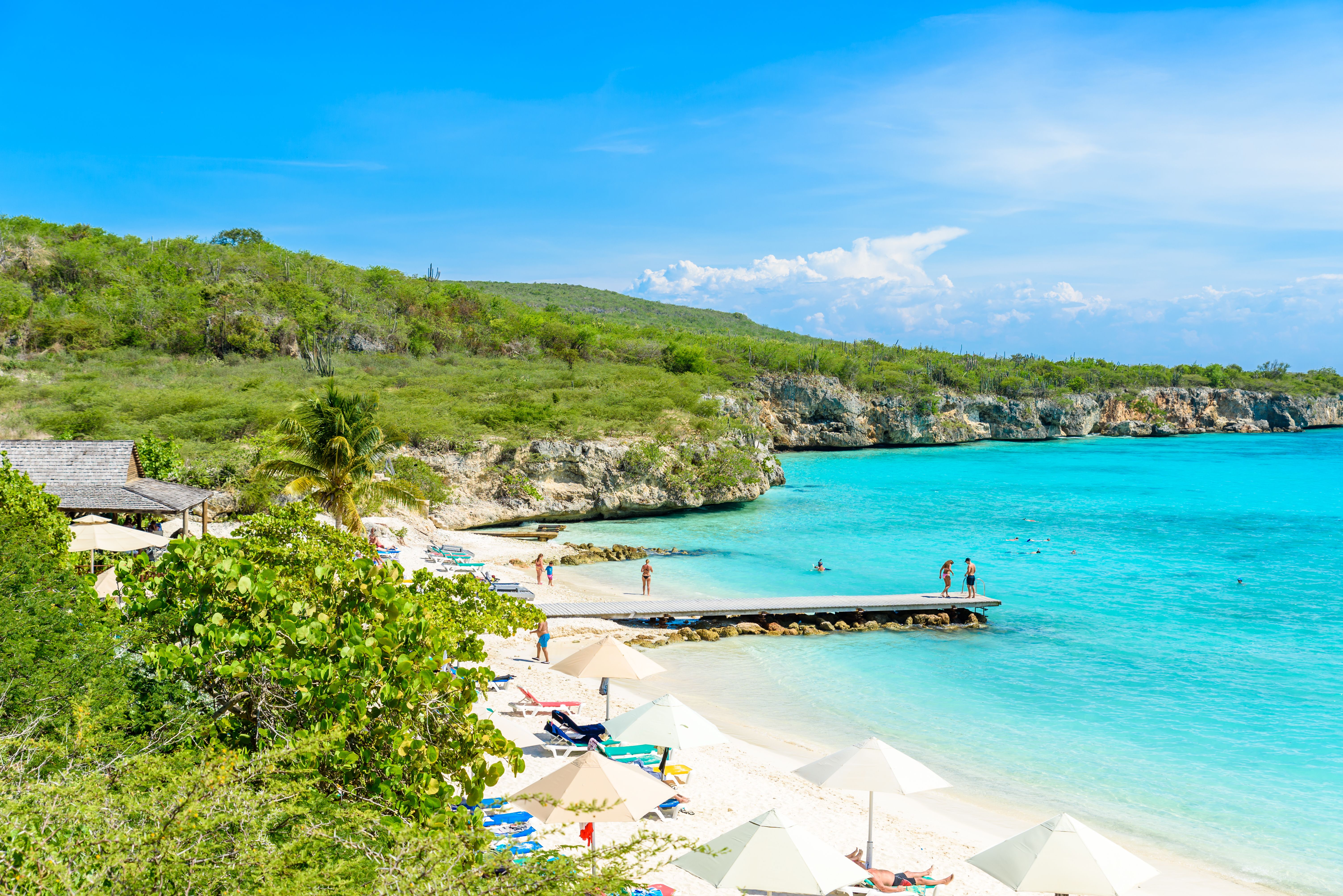 Scenic view of Playa Porto Mari in Curacao featuring white sandy beaches, turquoise waters, a wooden pier, sunbathers, and lush green hills in the background.