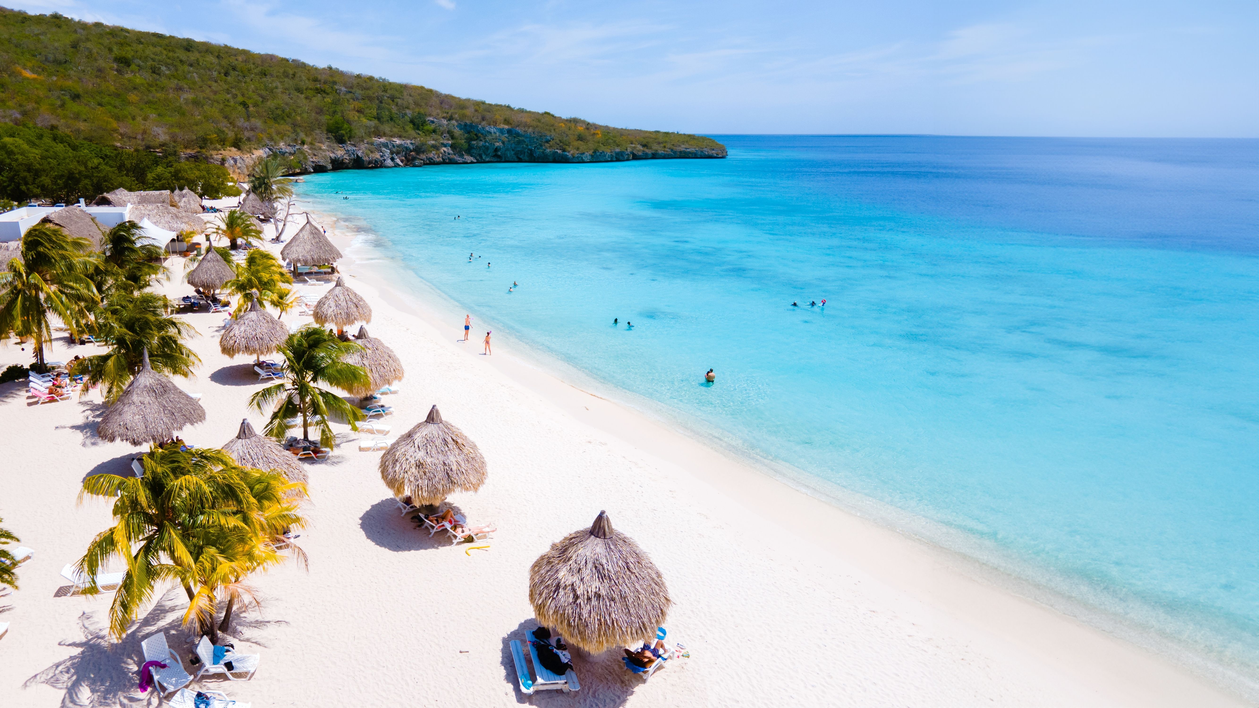 Breathtaking aerial view of Cas Abao Beach in Curacao with thatched umbrellas, swaying palm trees, white sand, and swimmers in clear blue waters.