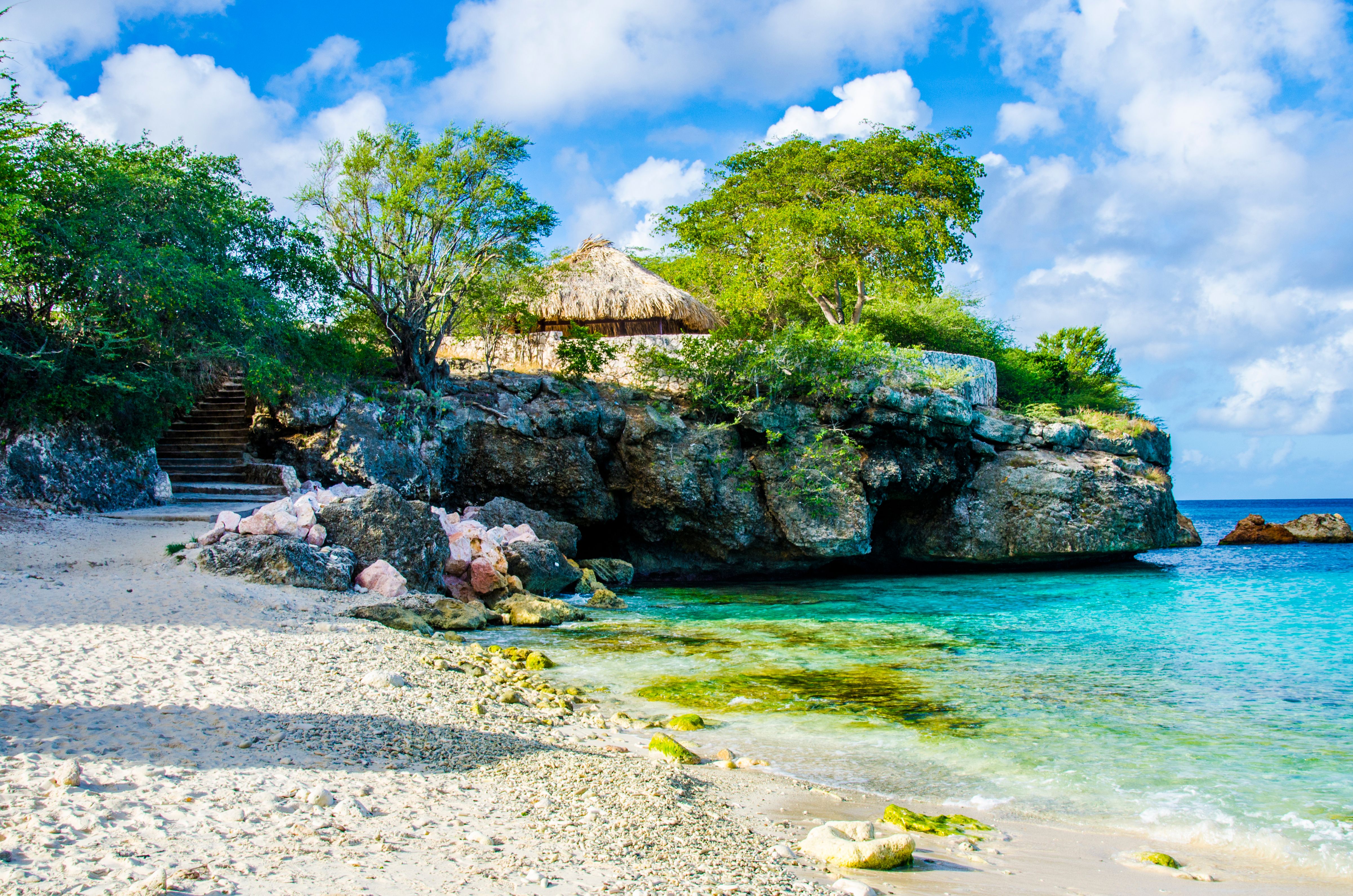 Grote Knip Beach in Curacao featuring a secluded cove with crystal-clear waters, a rocky ledge, a thatched gazebo, and a wooden staircase leading down to the beach.