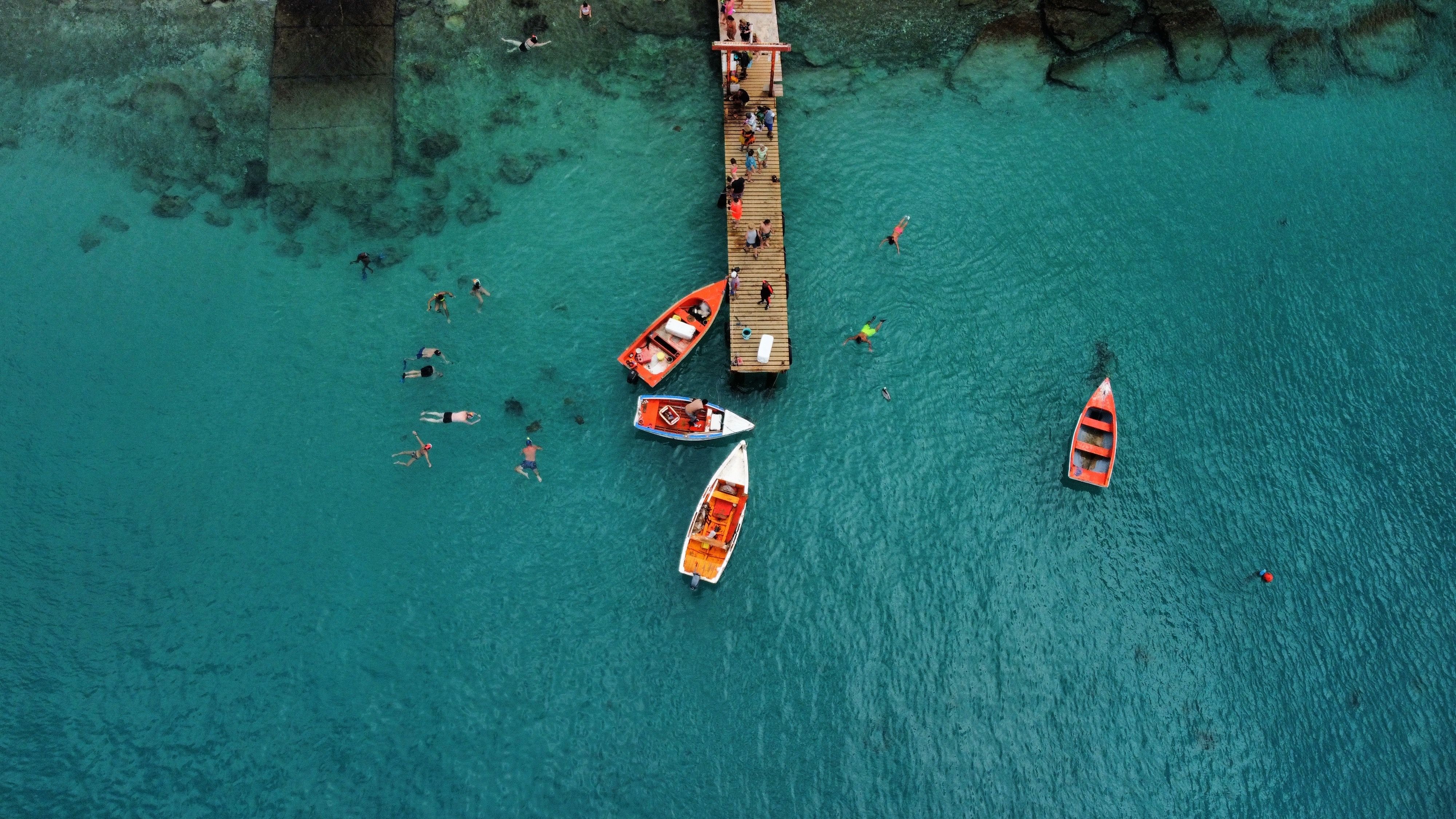 Aerial view of Playa Piskado in Curacao, with tourists swimming near colorful boats by a wooden pier, clear teal waters revealing underwater details.