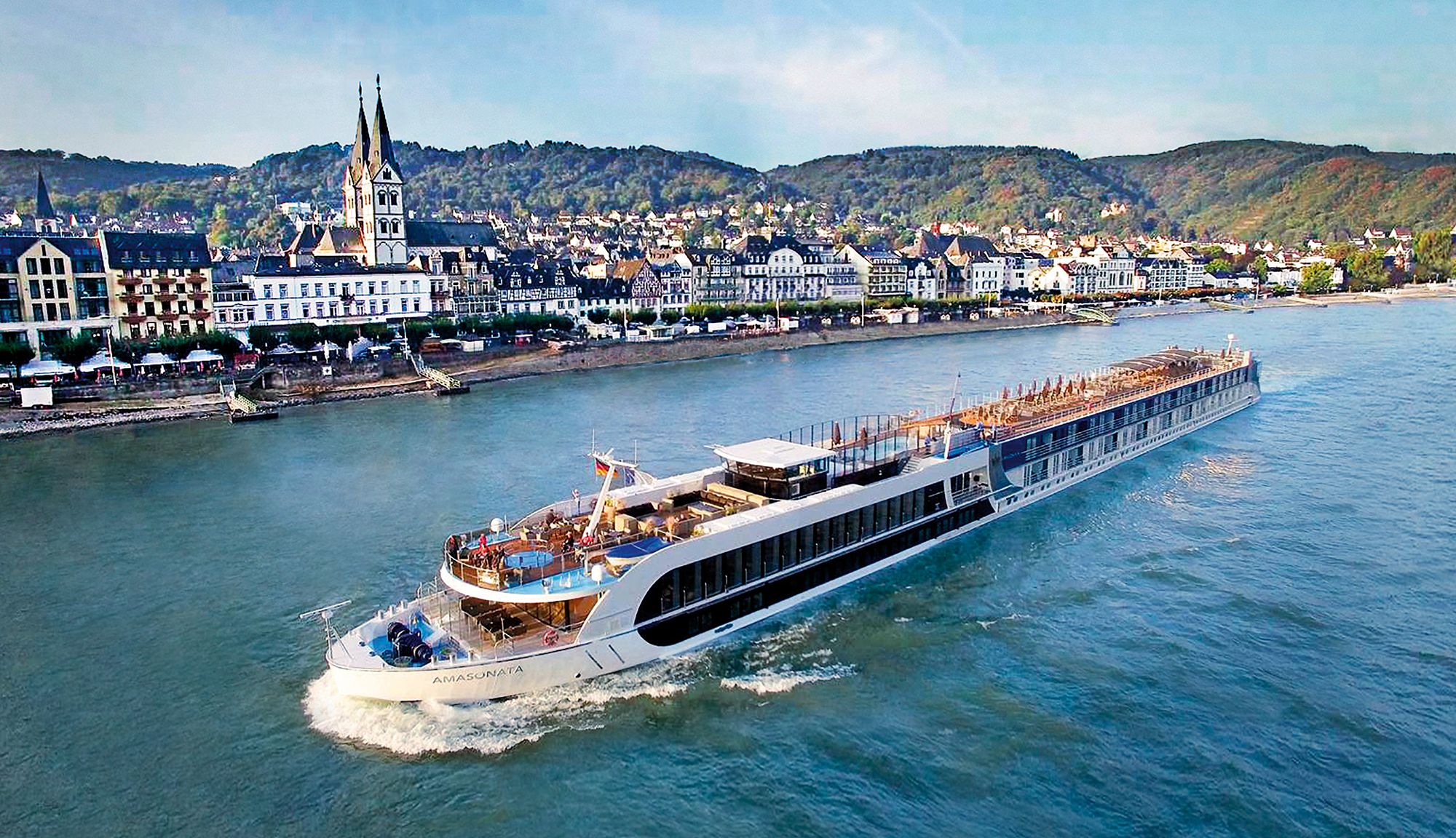 AmaSonata river cruise ship cruising on the Rhine River, passing by picturesque European architecture and lush green hills in the background.
