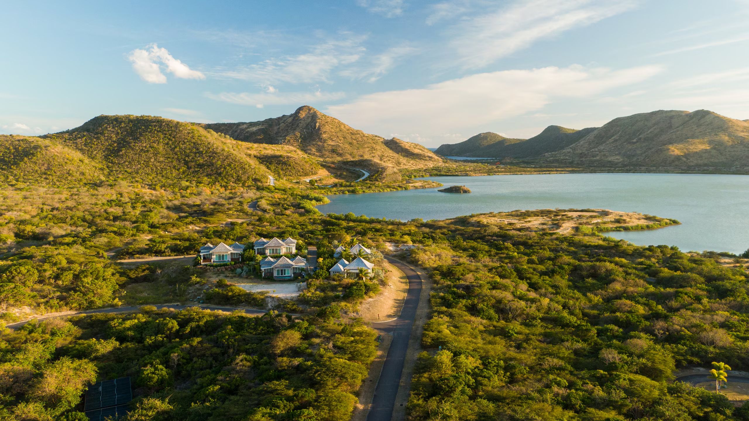Aerial view of resort bungalows amidst greenery, near a calm lake and winding roads with mountainous backdrop.