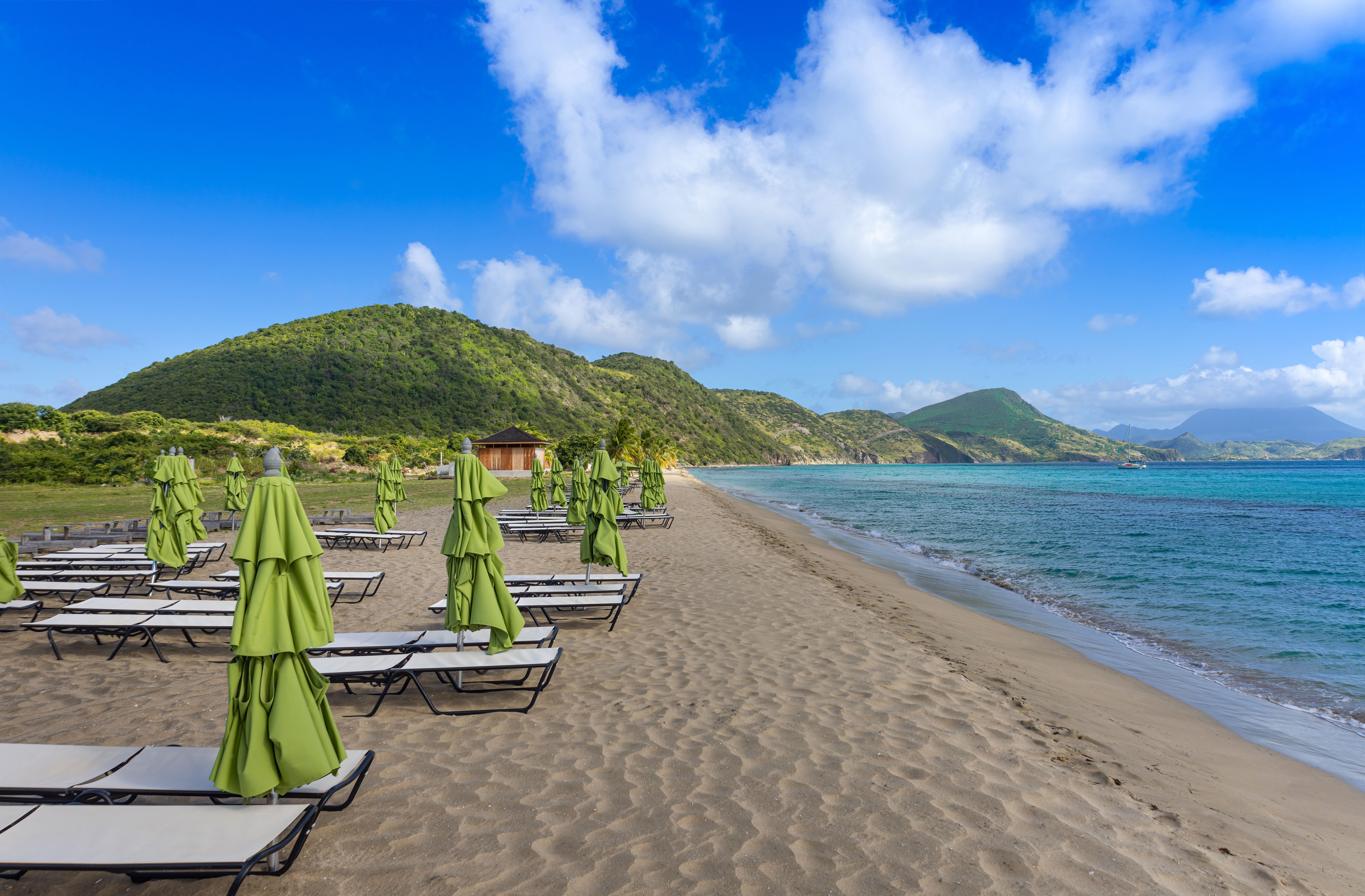Serene beach scene with rows of closed green umbrellas and lounge chairs on sandy shores framed by green hills.