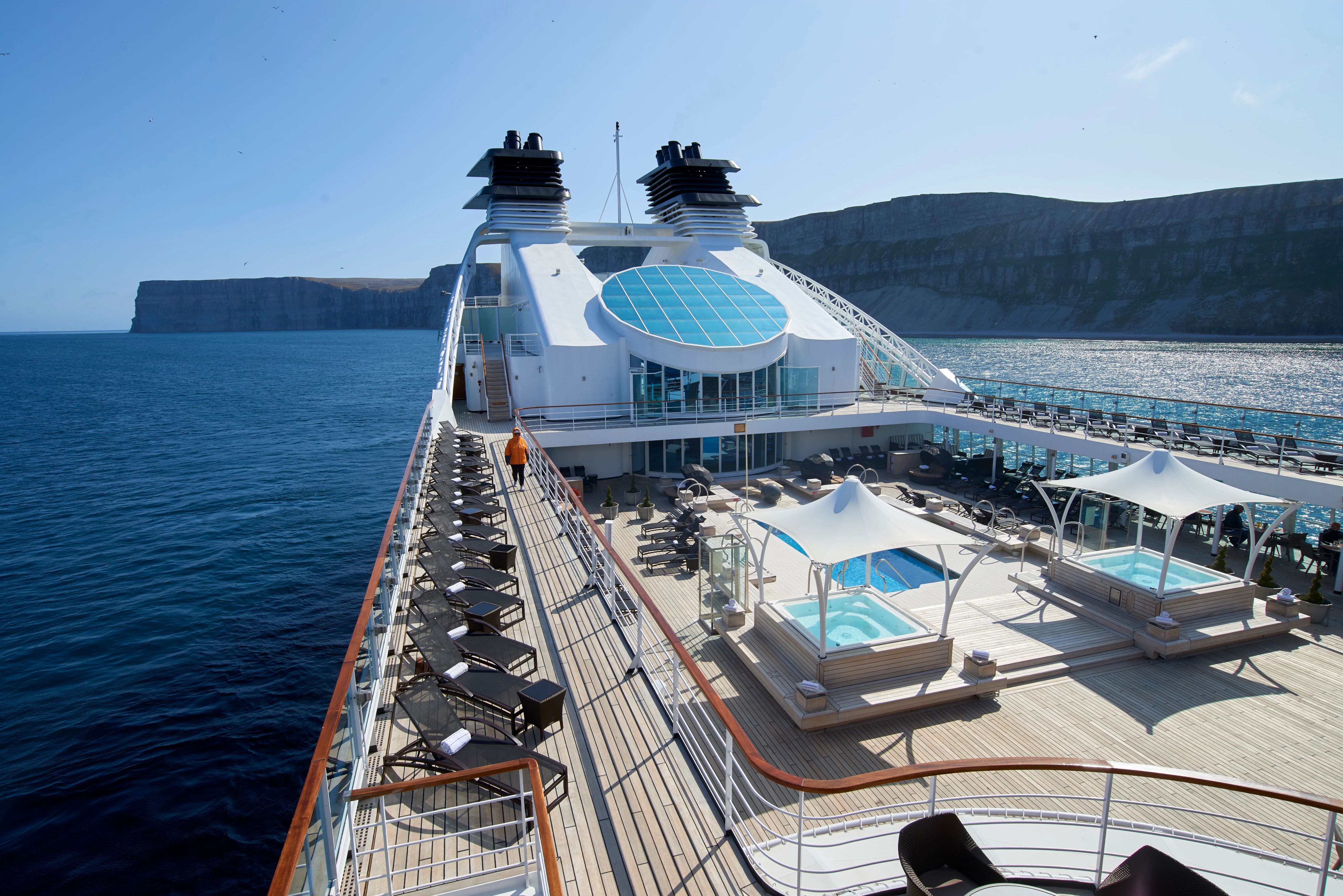 A luxurious pool deck on a Seabourn cruise ship with sun loungers and hot tubs, set against the stunning cliffs of an Alaskan fjord.