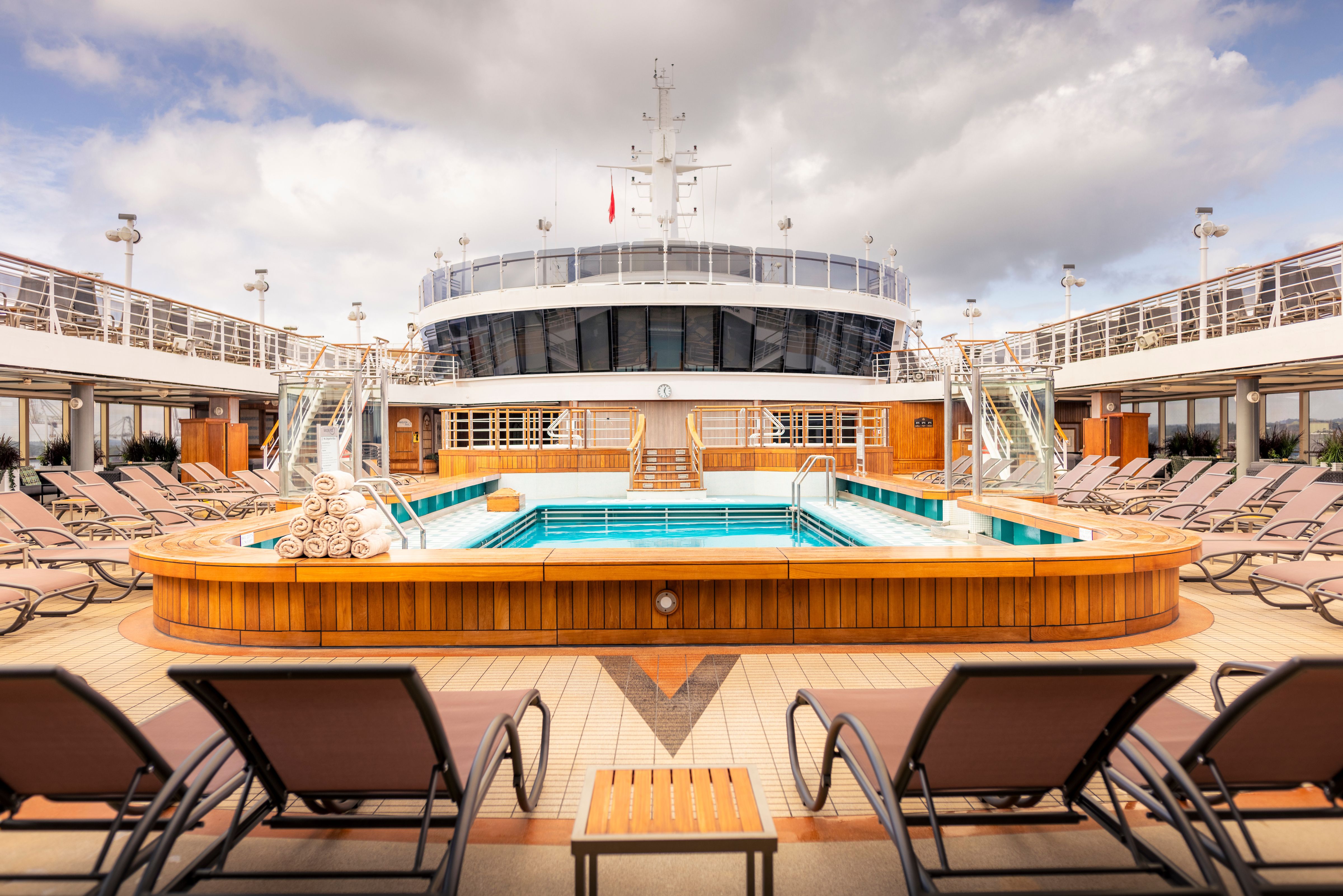 Pavilion pool area on Cunard Queen Victoria with loungers, towels, and a central pool under a partly cloudy sky.