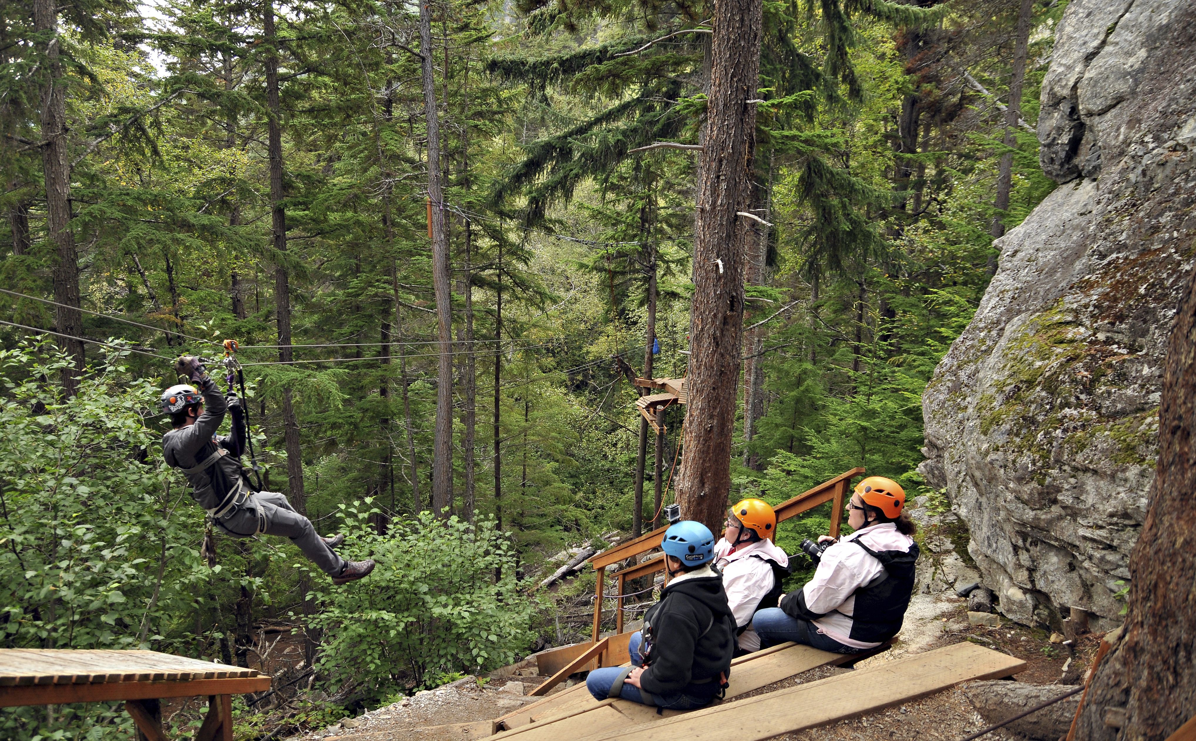 Ziplining in Alaska with lush green forest background, three onlookers in helmets. 