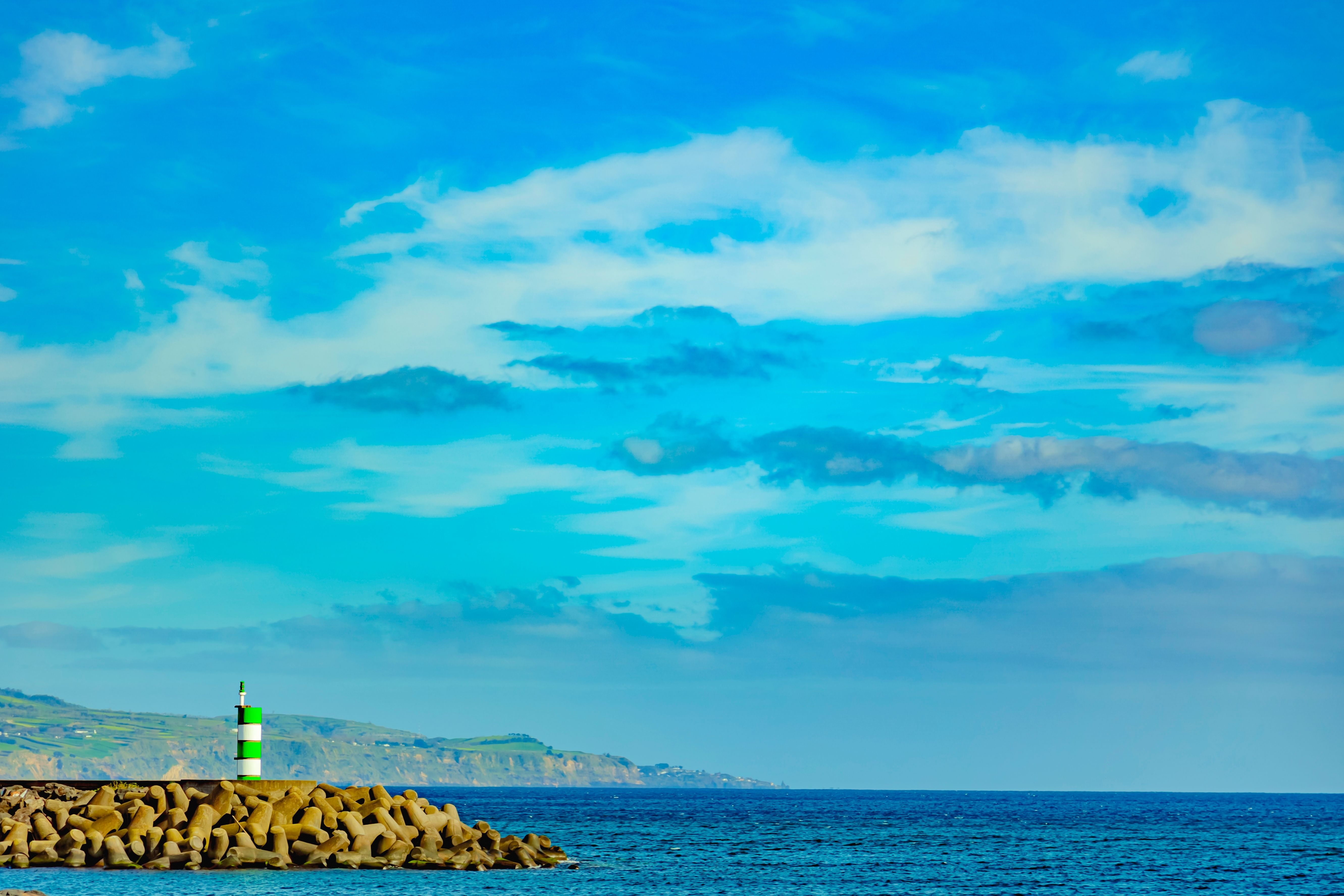 Scenic view of Ponta Delgada coastline with a small green and white lighthouse, rocky breakwater, and blue ocean, Azores.