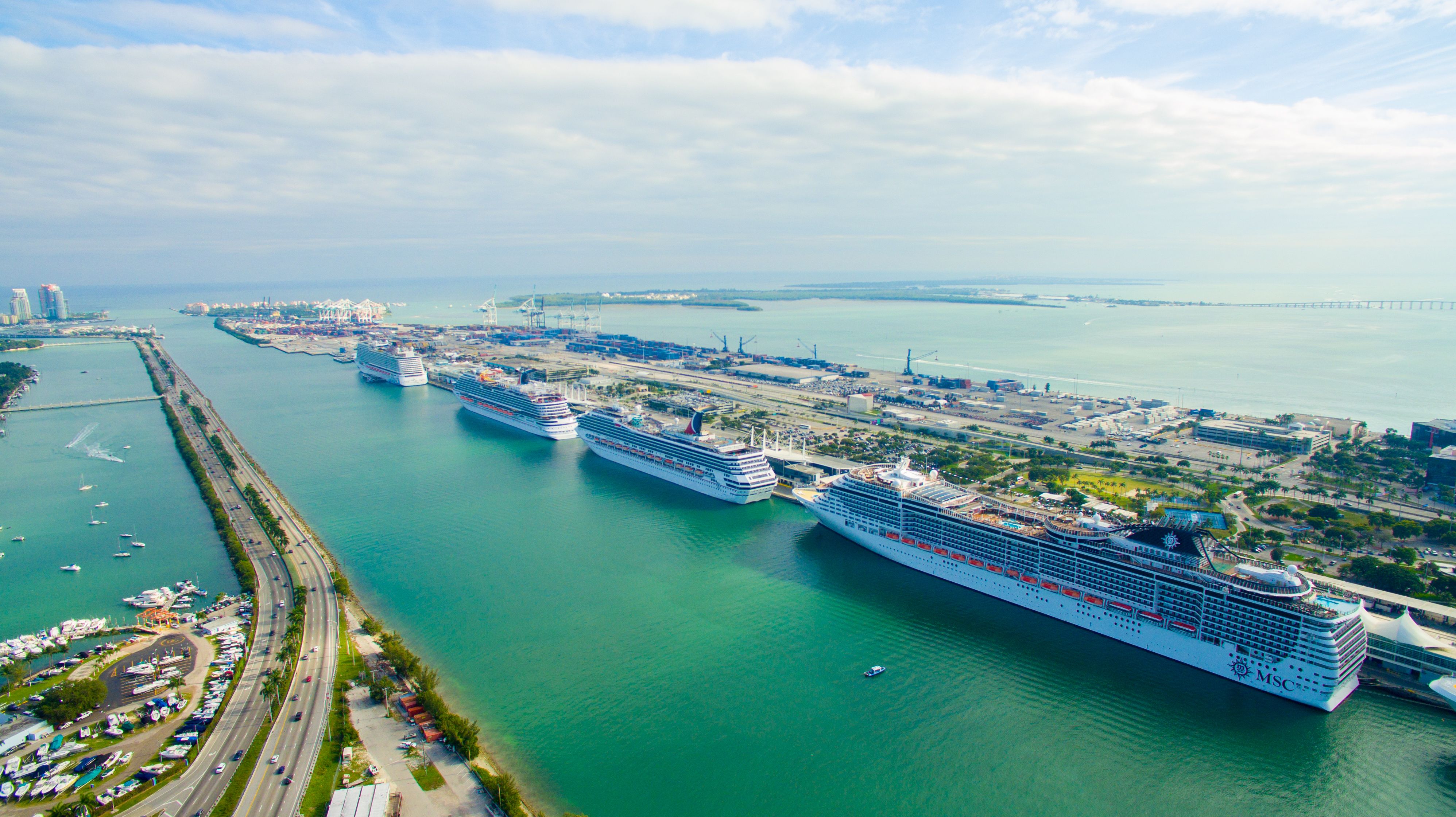 Multiple cruise ships docked at PortMiami, with the city's skyline and busy port facilities in the background, under a partly cloudy sky.
