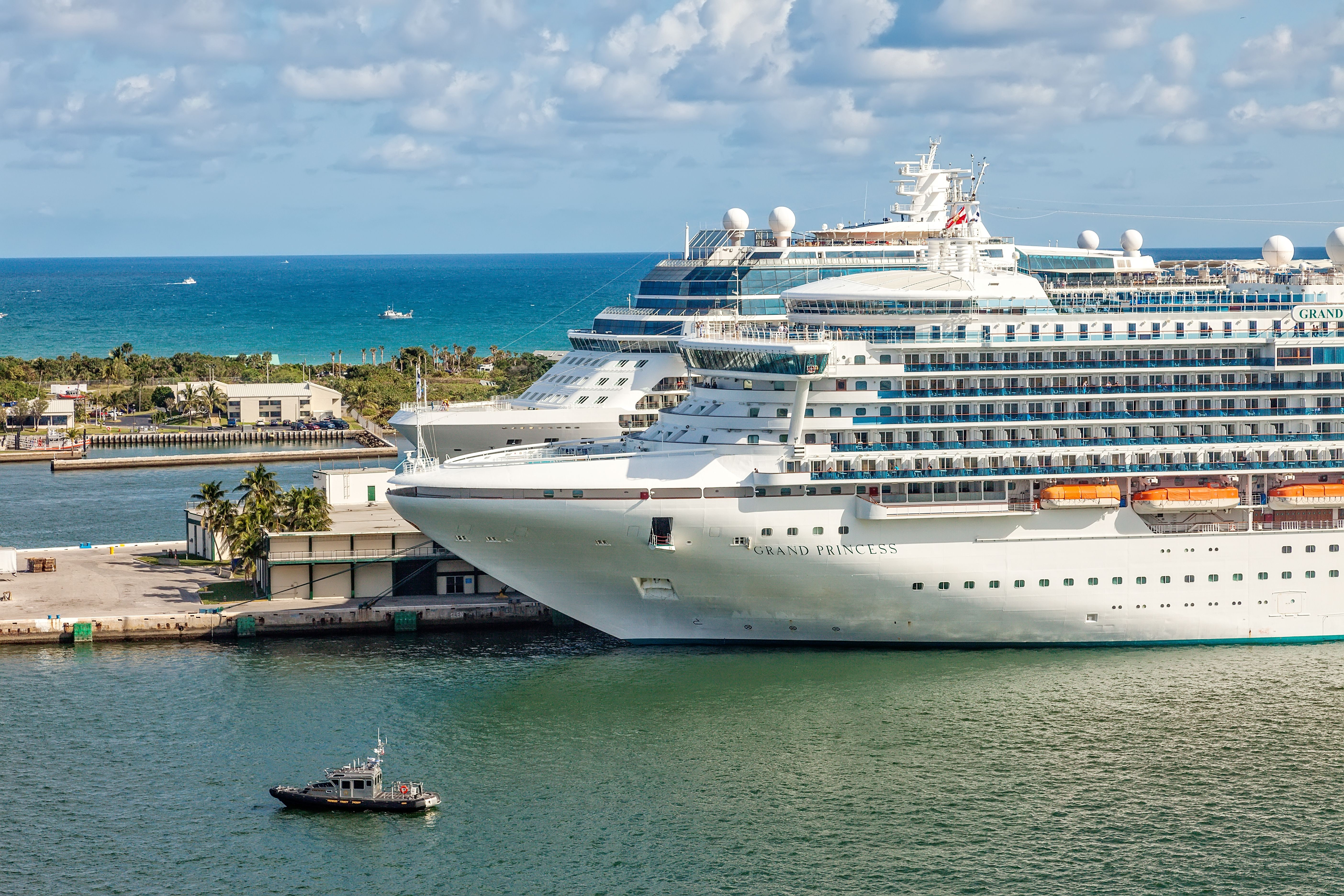 The Grand Princess docked at Port Everglades in Fort Lauderdale, Florida, with a small tugboat nearby and the ocean in the background.