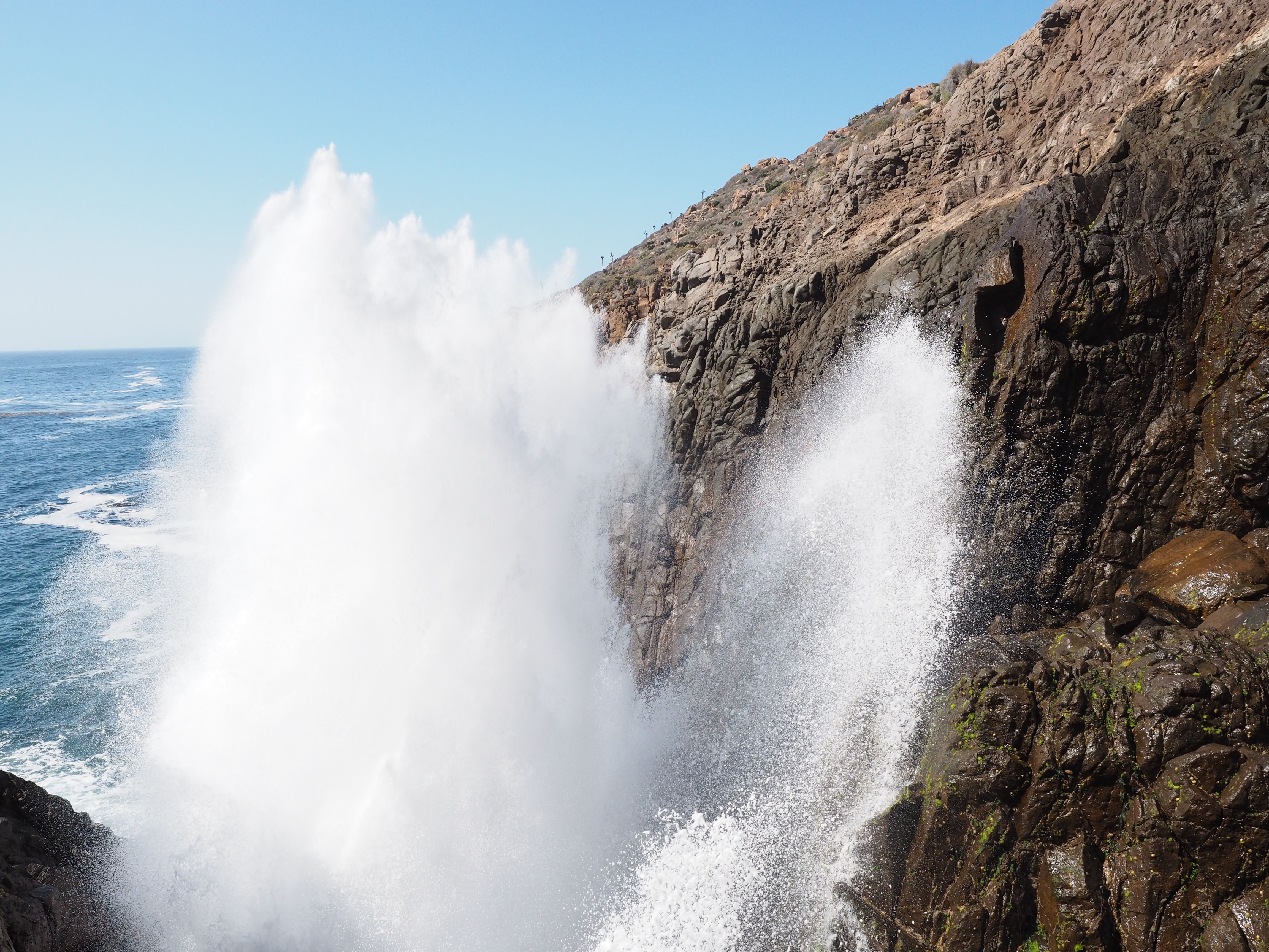 Powerful ocean geyser La Bufadora shooting water up rocky cliff, creating mist under a clear blue sky. Popular natural attraction near Ensenada, Mexico.