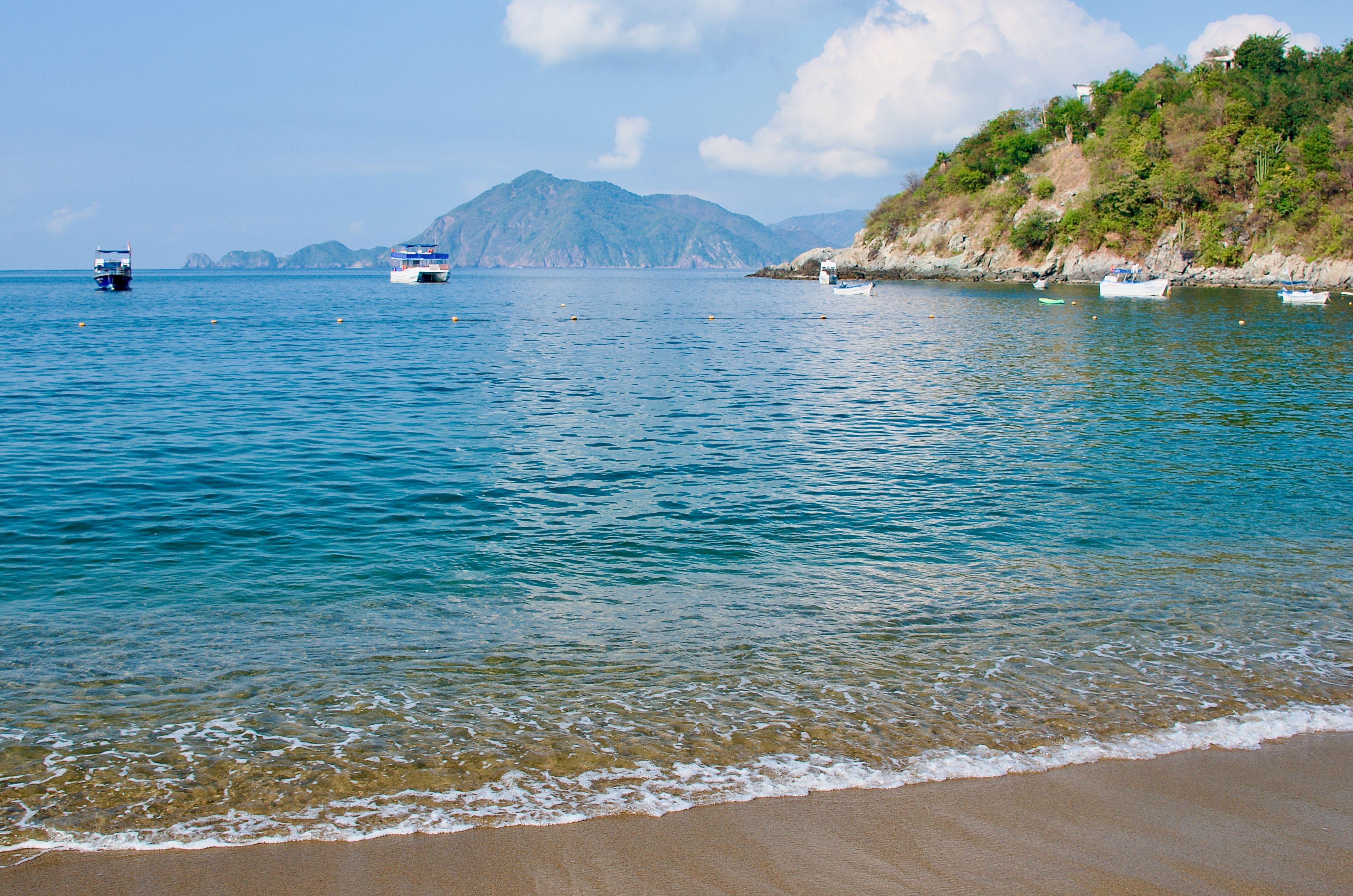 Scenic view of Manzanillo shore with calm blue water, anchored boats, and lush green hills under a clear sky.