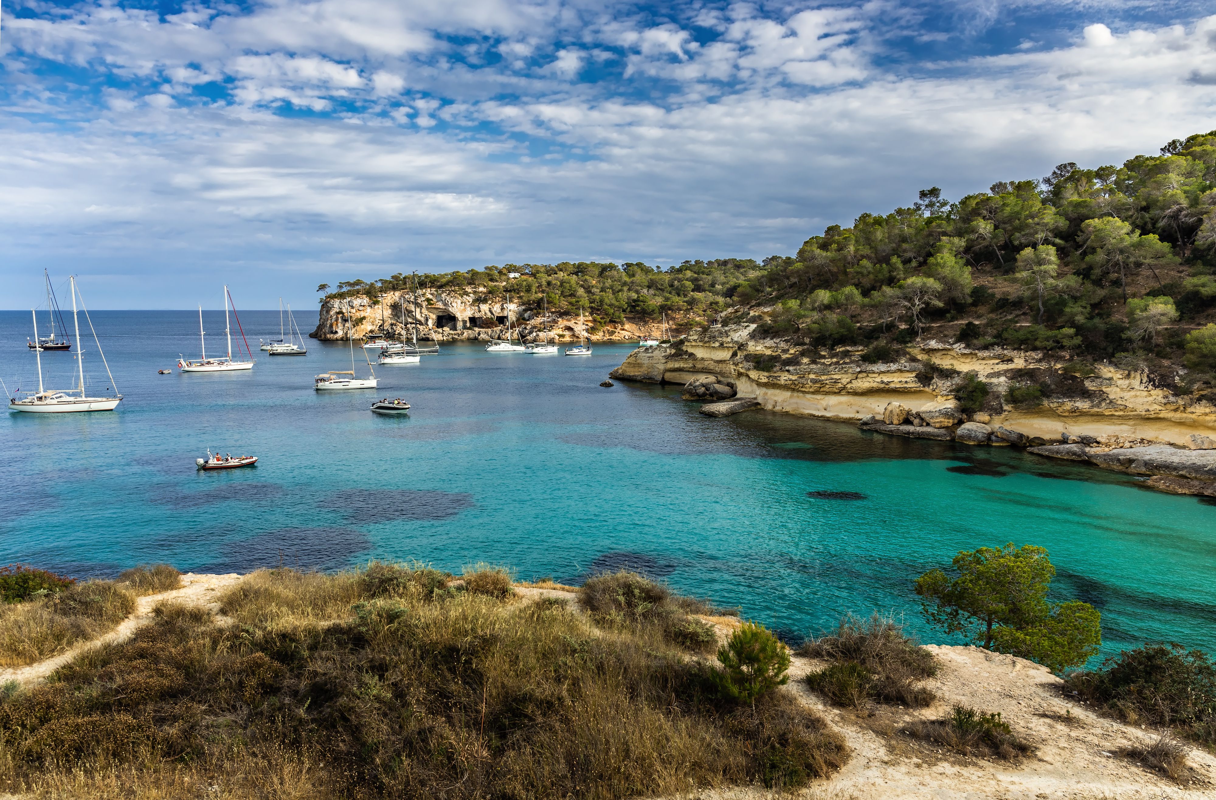 Cala del Mago cove with anchored sailboats, clear turquoise waters, and rugged cliffs in Mallorca, Spain.