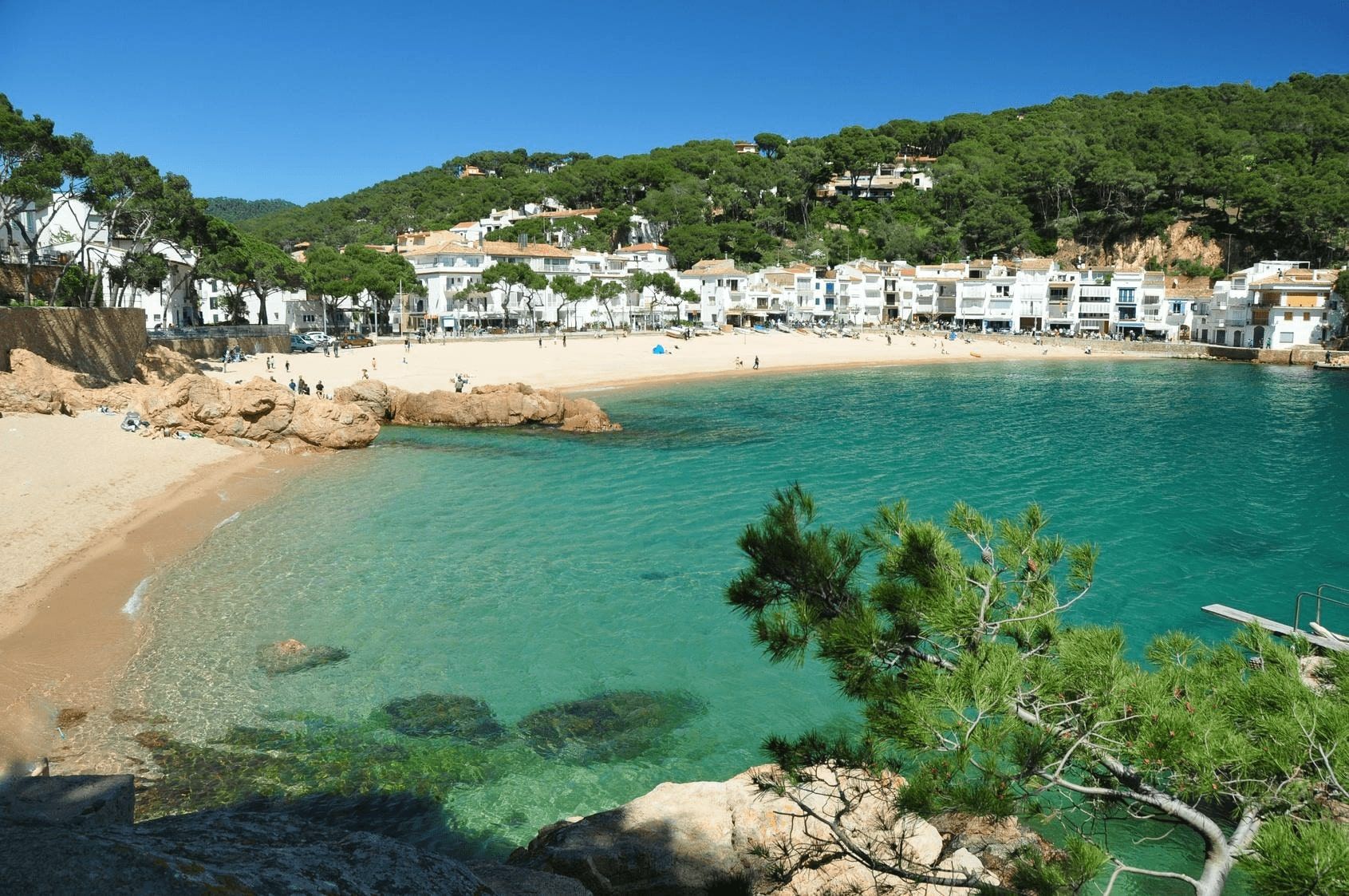 Scenic Cala Balva beach with turquoise waters, sandy shore, and white houses nestled among lush green hills in Costa Brava, Spain