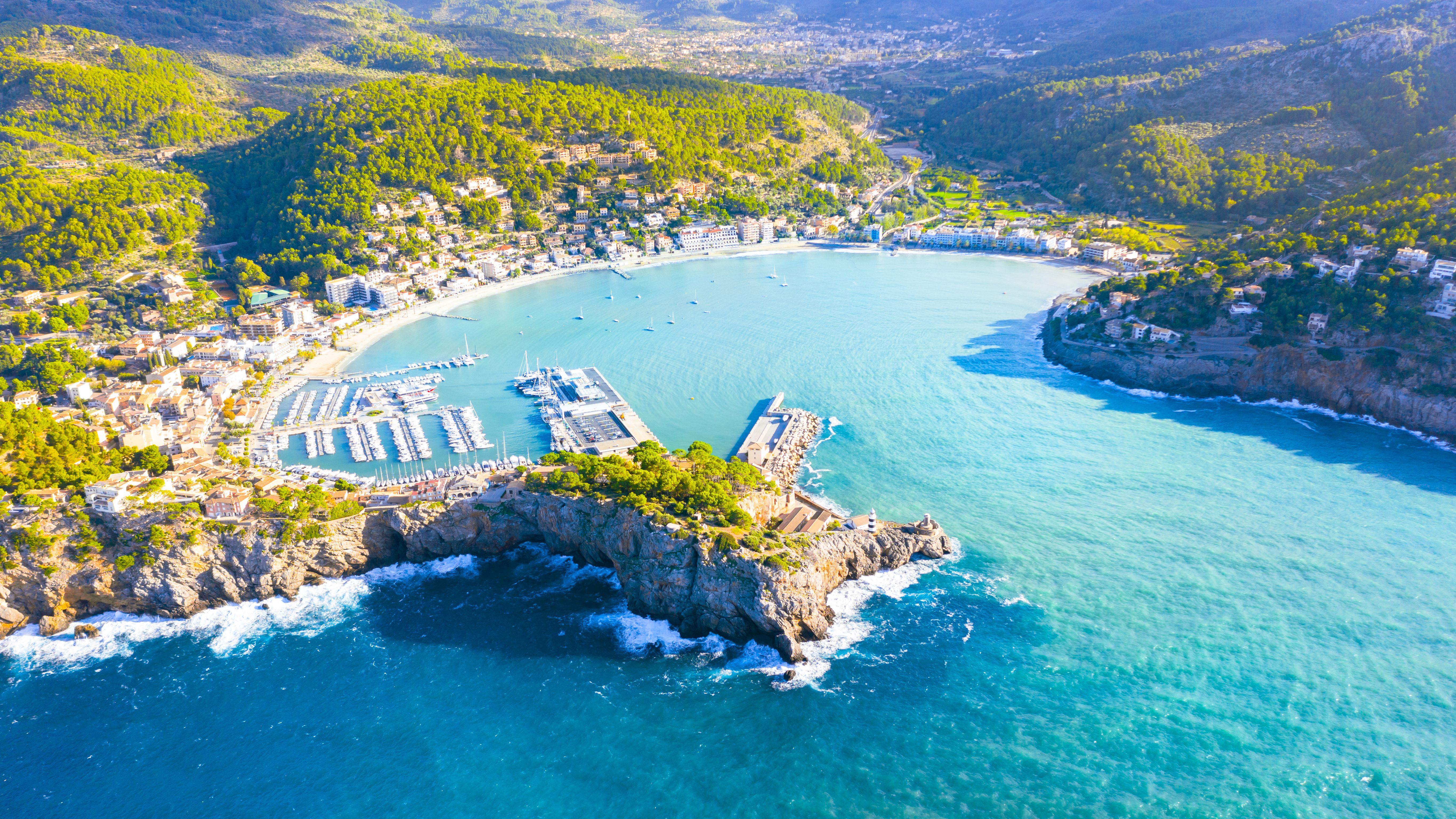 Aerial view of Port de Sóller in Mallorca, Spain, showcasing a picturesque bay, marina filled with boats, and surrounding green hills under a bright sky.