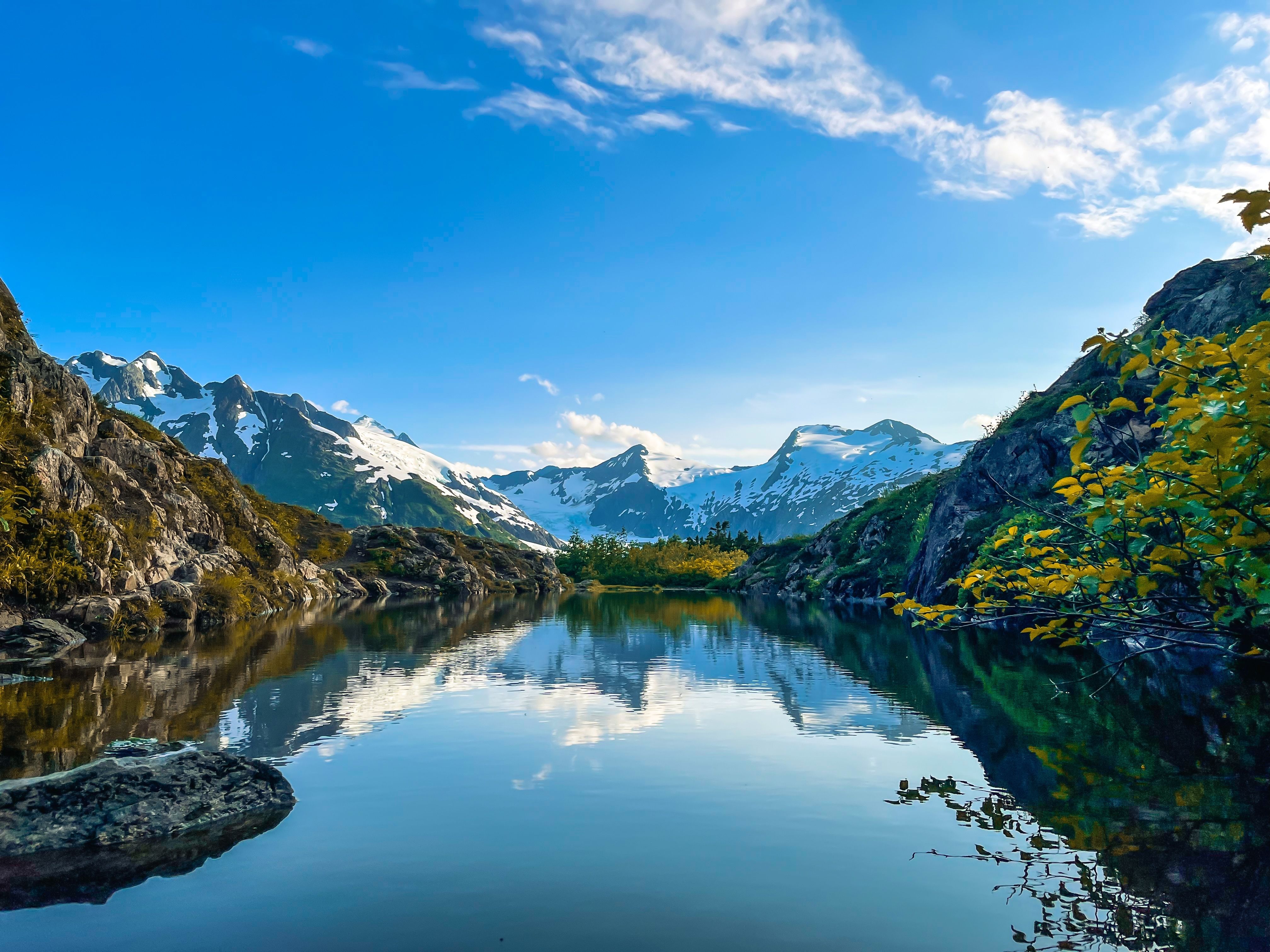 Scenic view of snow-capped mountains and a serene lake in Alaska, under a bright blue sky with scattered clouds.