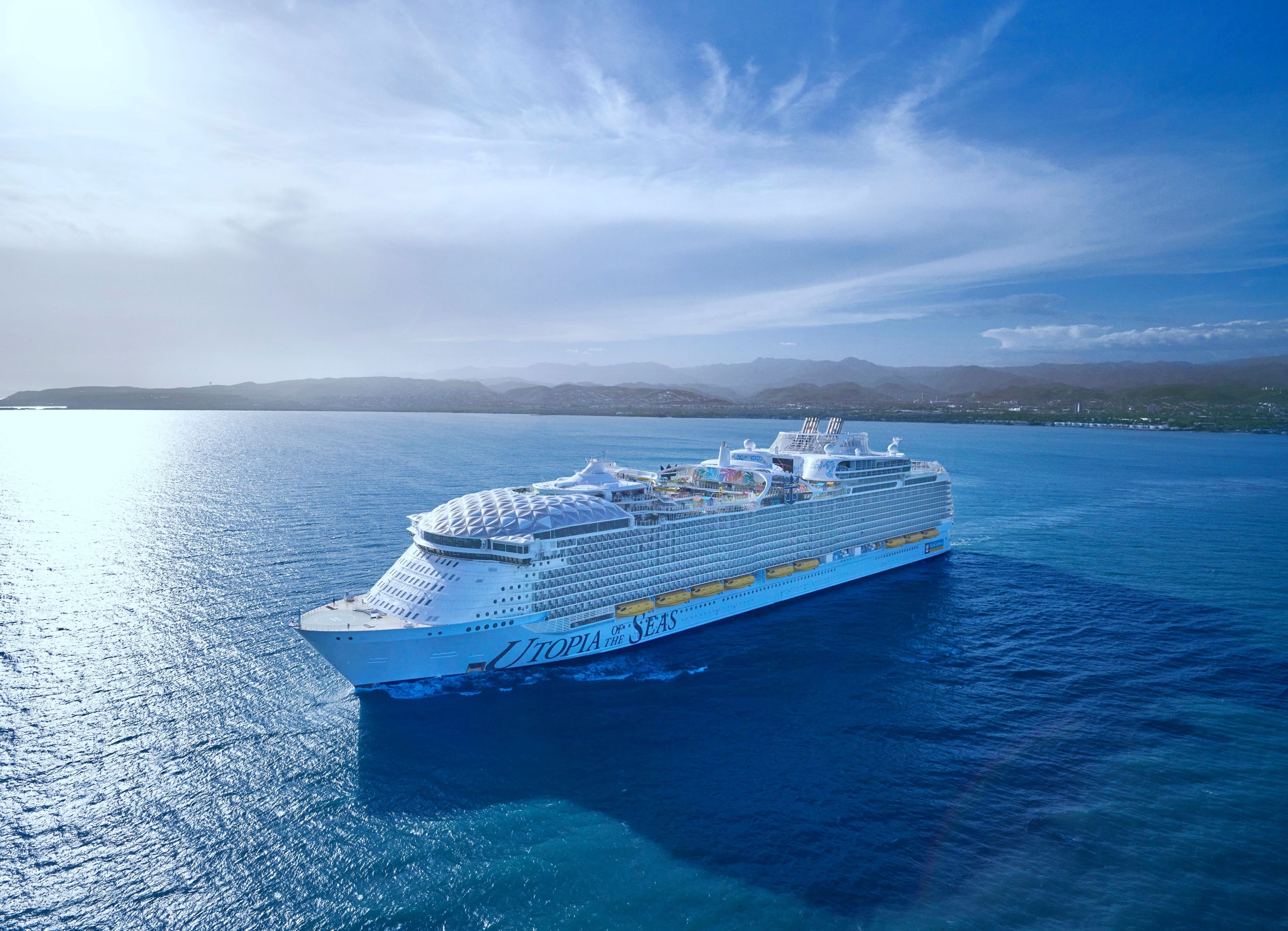 Aerial view of Royal Caribbean's Utopia of the Seas, a large cruise ship sailing in clear blue waters with coastal mountains in the background.