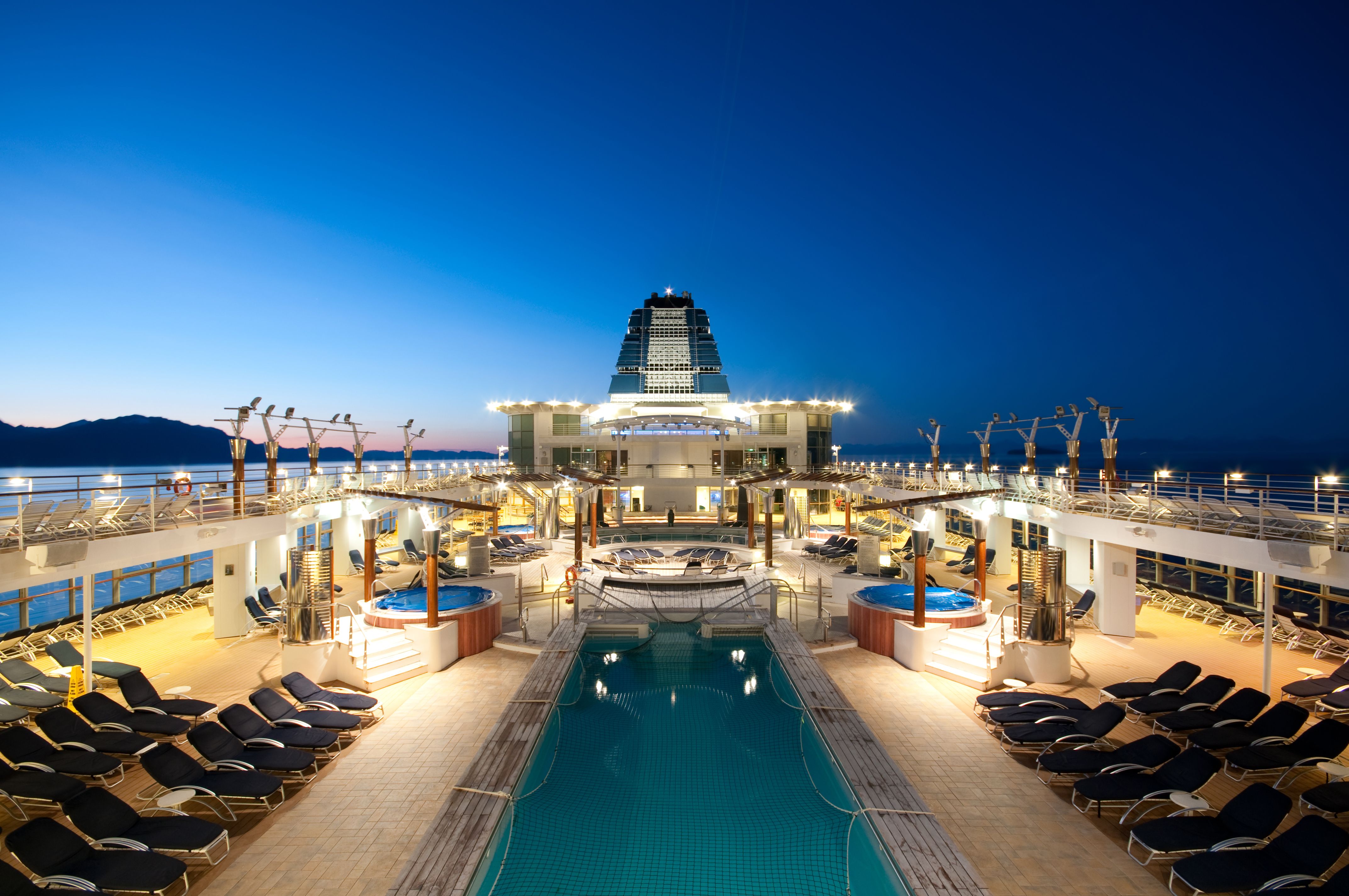 Luxurious cruise ship pool deck at sunset in Alaska, featuring sun loungers, pools, and jacuzzis with a scenic ocean backdrop.