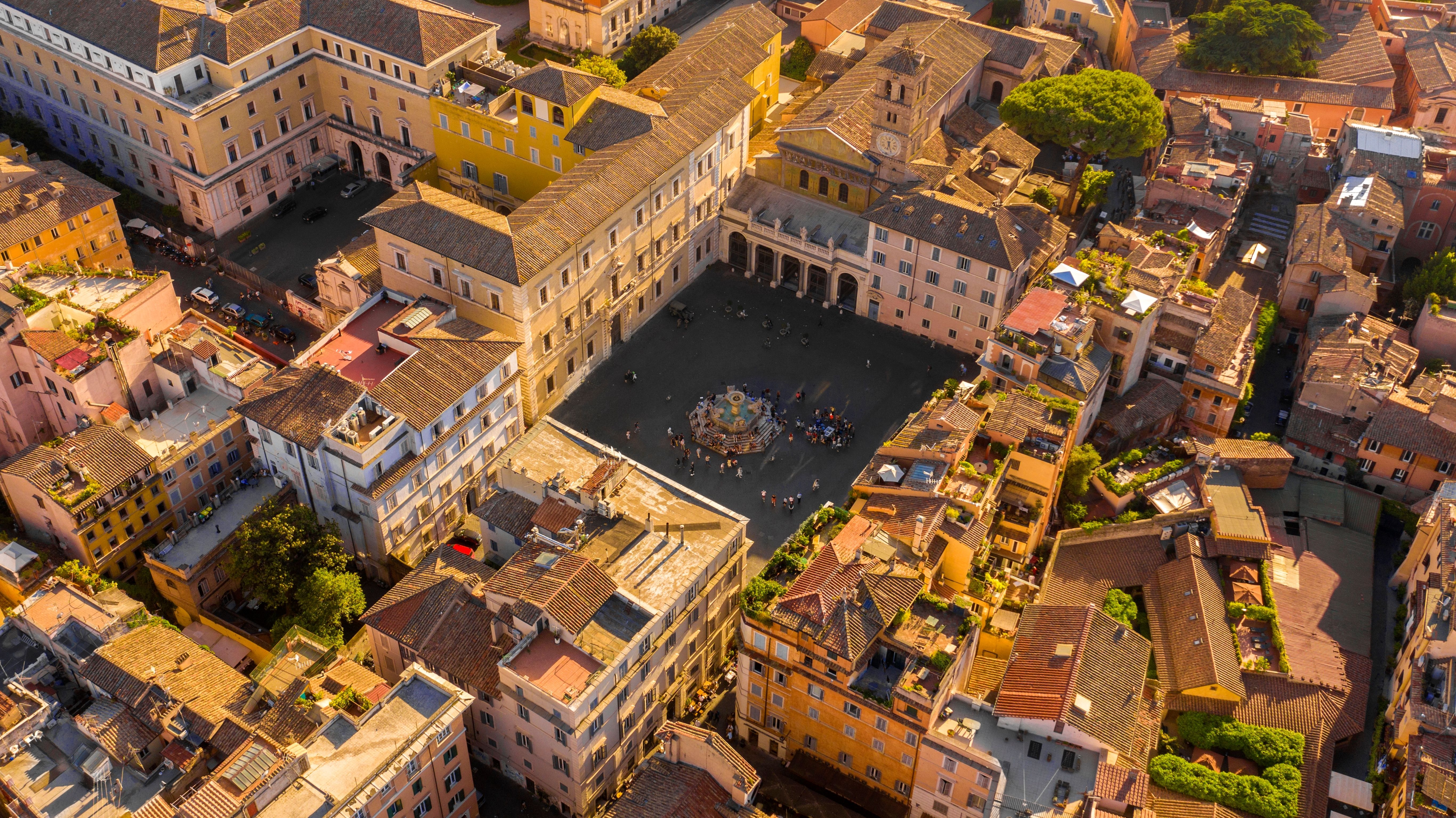 Aerial view of Trastevere, Rome, showcasing historic buildings, a central square with a fountain, and bustling streets, captured in warm, golden light. Ideal for Mediterranean cruise excursions.