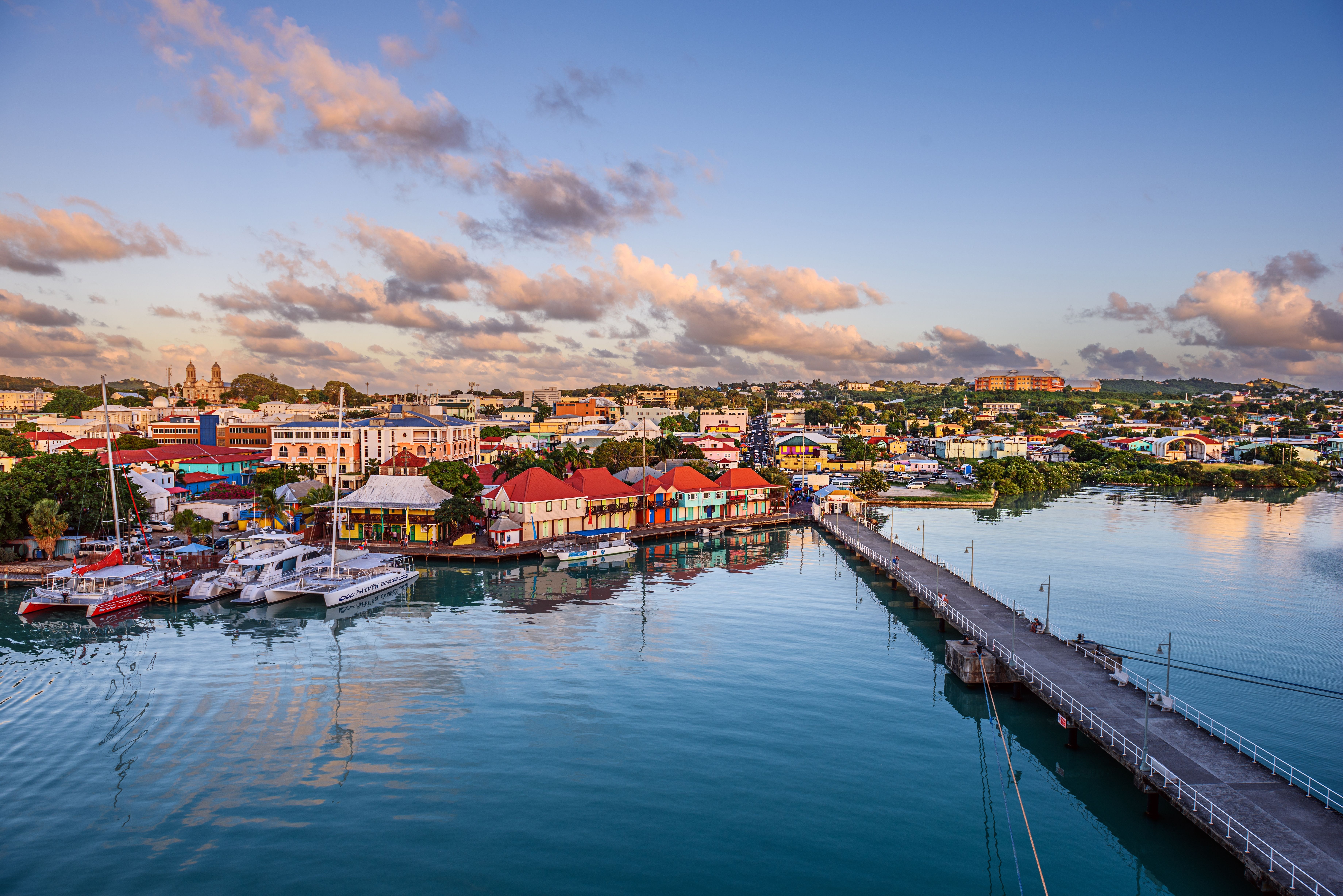 Aerial view of St. John's harbor in Antigua at sunset, showcasing colorful buildings, yachts, and a long pier reflected in the calm water.
