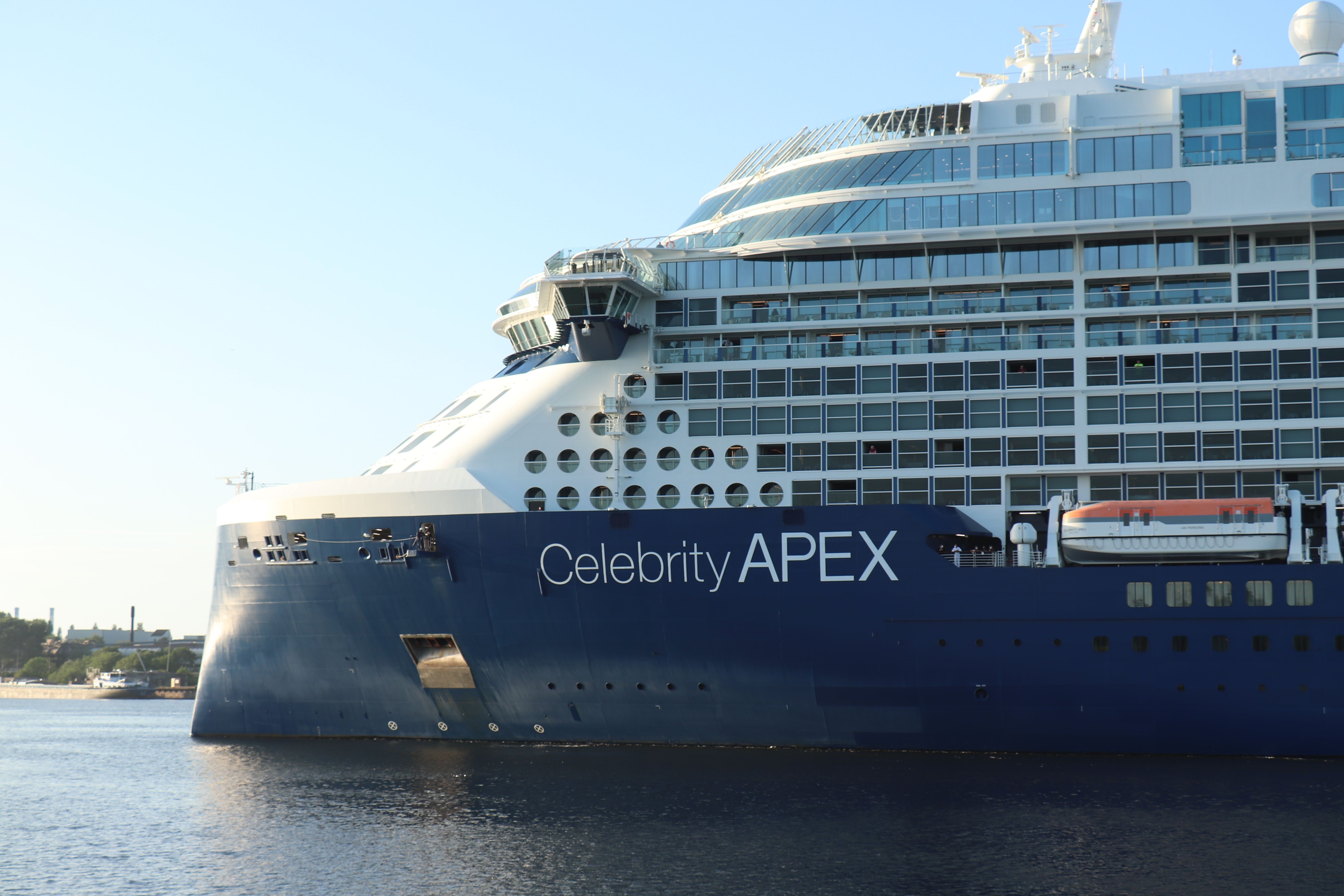 A close-up view of the Celebrity Apex cruise ship docked at a port during daylight. The ship’s name is prominently displayed, with the ship's modern and elegant design visible against a calm water backdrop.