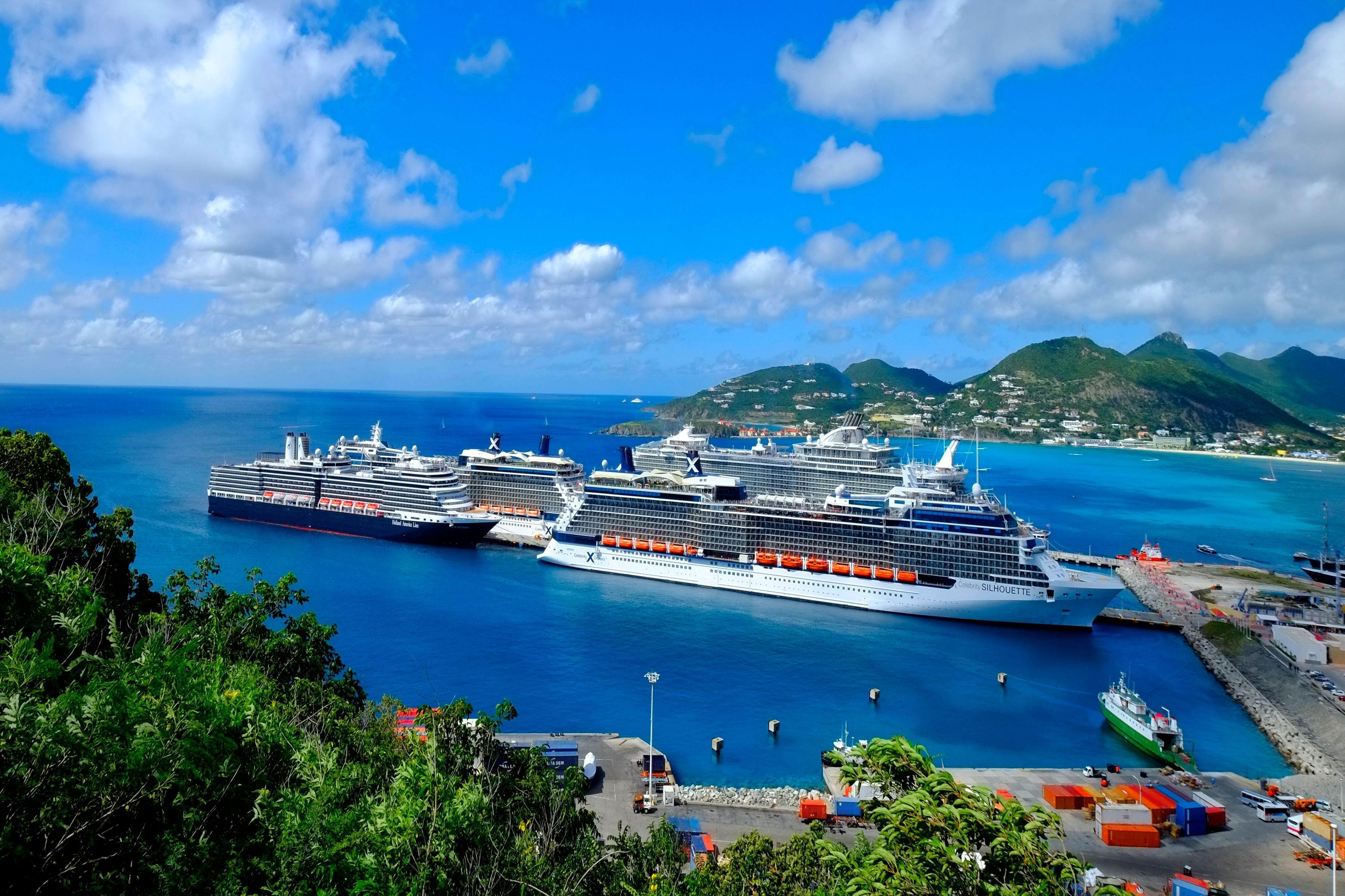 An aerial view of the Celebrity Silhouette cruise ship docked alongside other cruise ships in a picturesque Caribbean port. The scene features lush green hills and bright blue waters, typical of a tropical paradise.