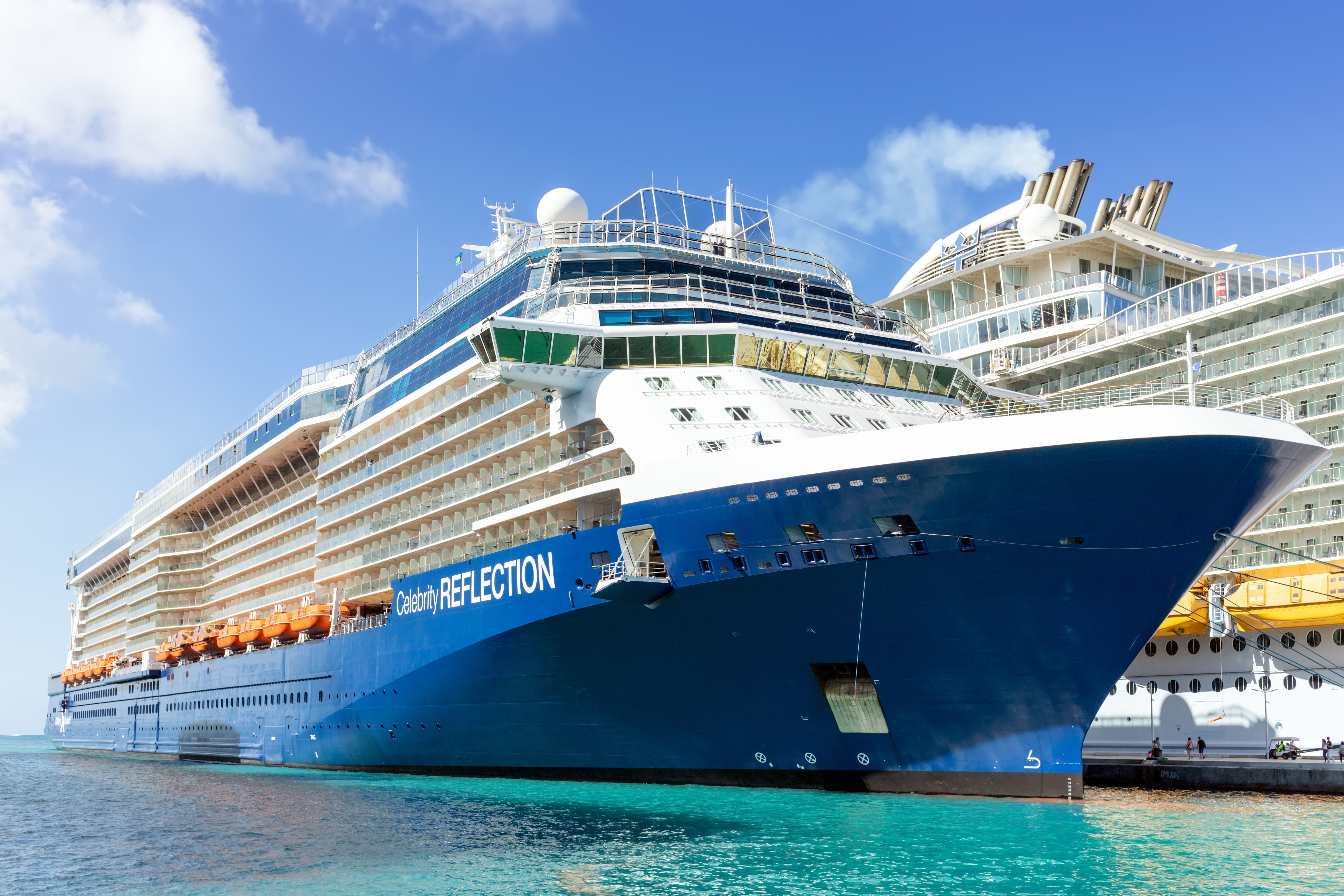 A stunning daytime view of the Celebrity Reflection cruise ship docked at port. The ship's sleek design and multiple balconies are visible, with a bright blue sky and calm turquoise waters enhancing the scene.