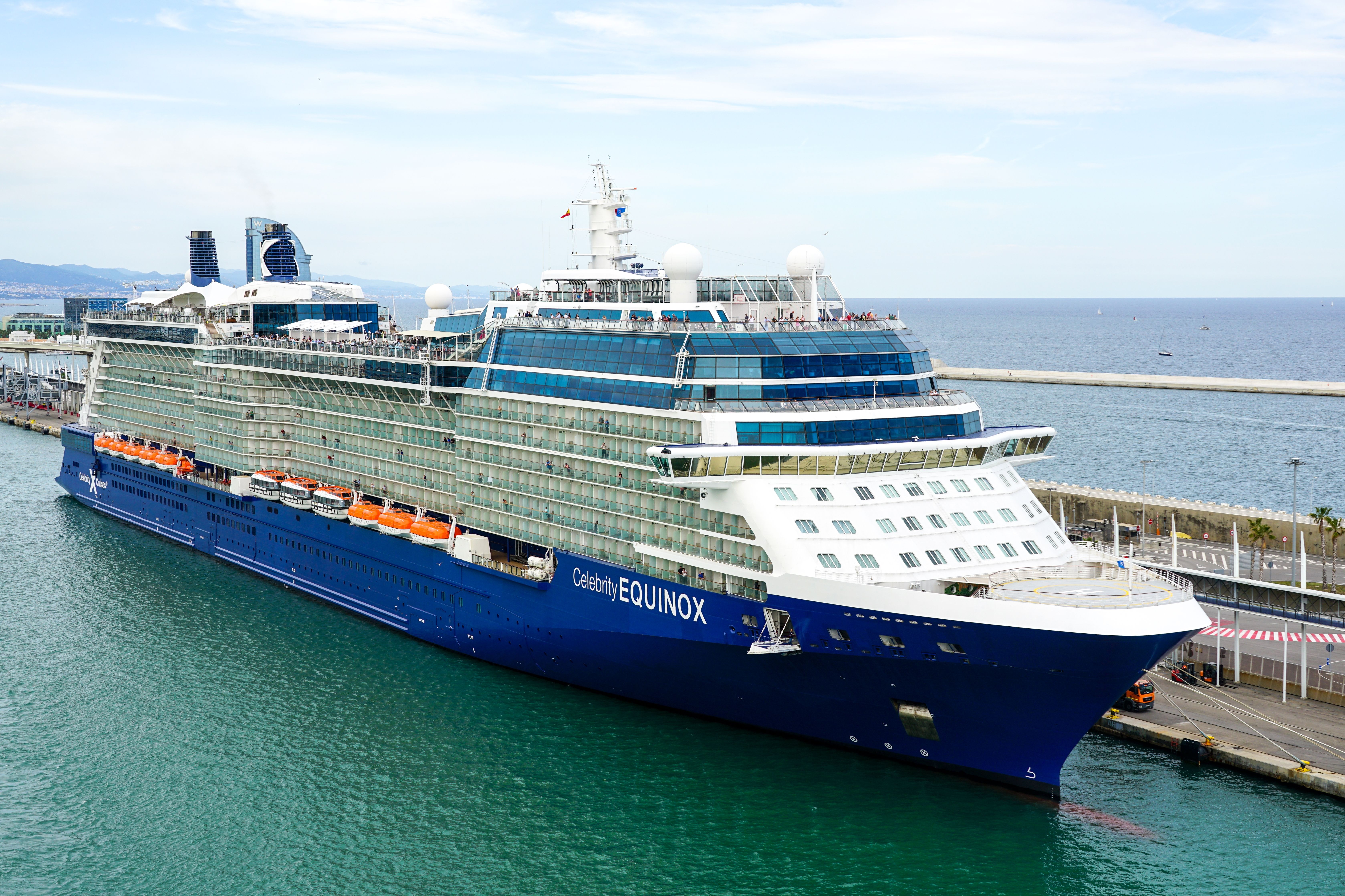 A daytime view of the Celebrity Equinox cruise ship docked at a busy port. The ship's sleek, modern lines and extensive balcony spaces are clearly visible, with a calm sea surrounding the vessel.