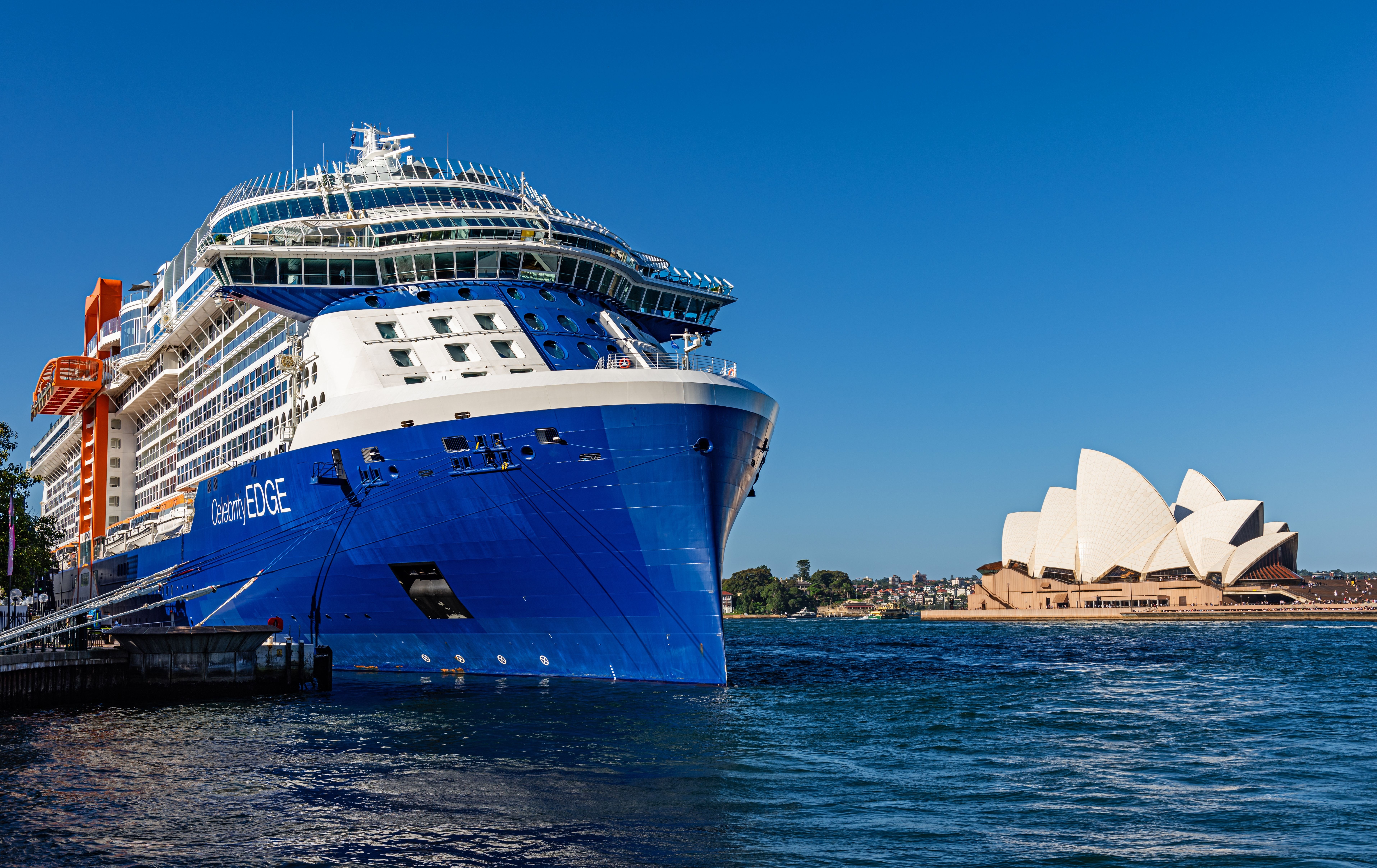The Celebrity Edge cruise ship docked at the Sydney Harbor, with the iconic Sydney Opera House visible in the background. The ship's modern design is highlighted by a clear blue sky and the serene harbor waters.