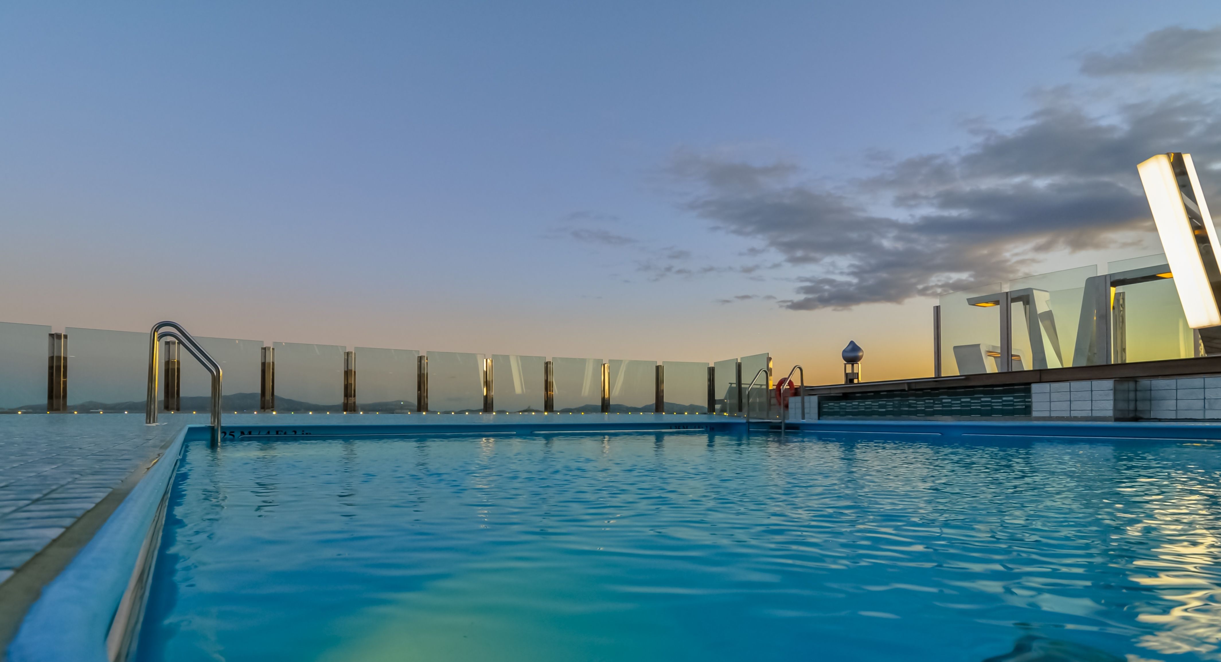 A serene infinity pool on a luxury cruise ship at sunset. The pool is surrounded by glass panels, offering uninterrupted views of the horizon as the sky takes on hues of purple and gold.