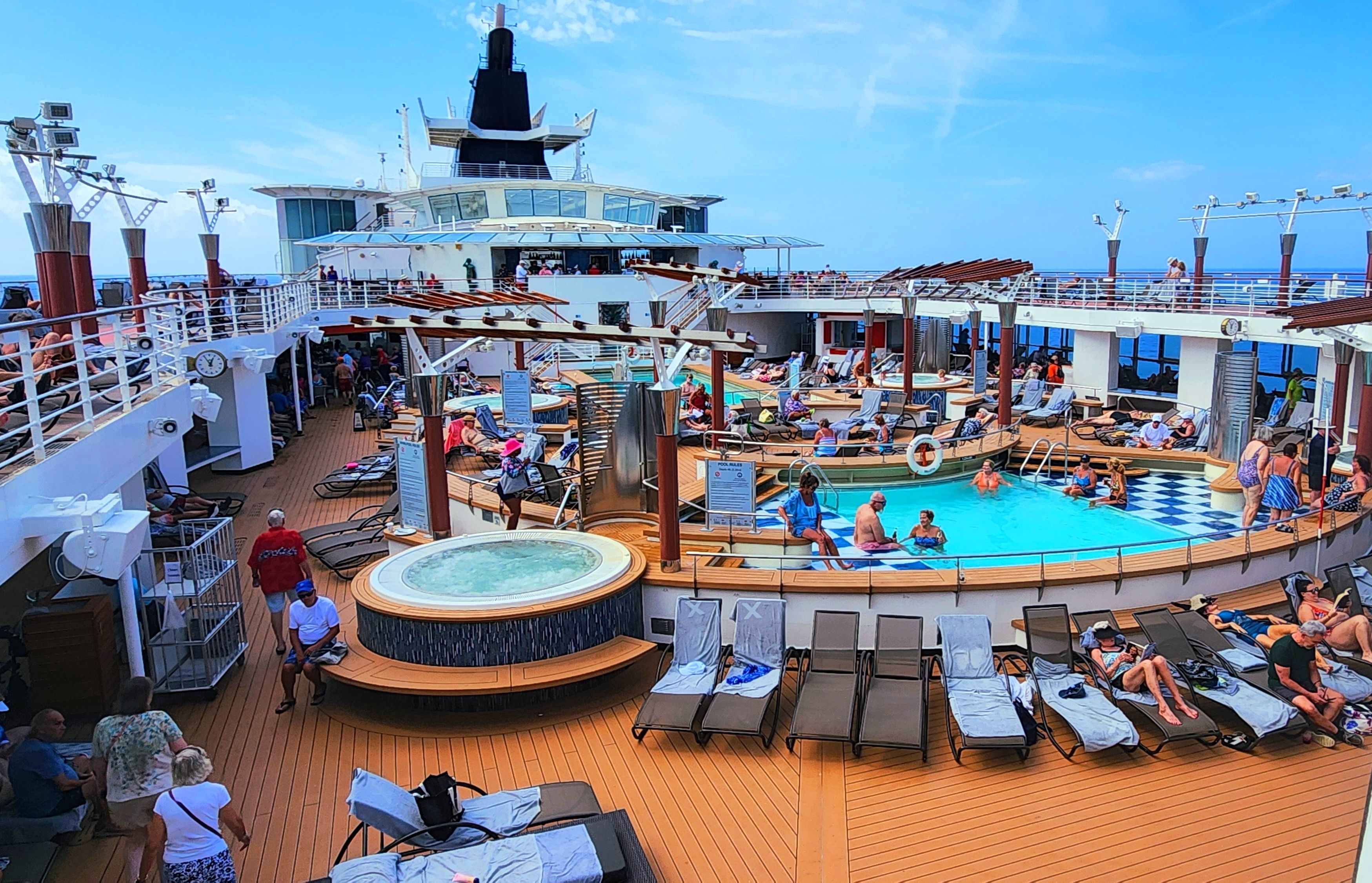 A vibrant pool deck on a Celebrity cruise ship. Passengers lounge on sunbeds around a central pool and jacuzzi, enjoying the relaxed ambiance. The deck is adorned with modern design elements, offering a stylish and comfortable setting for relaxation.