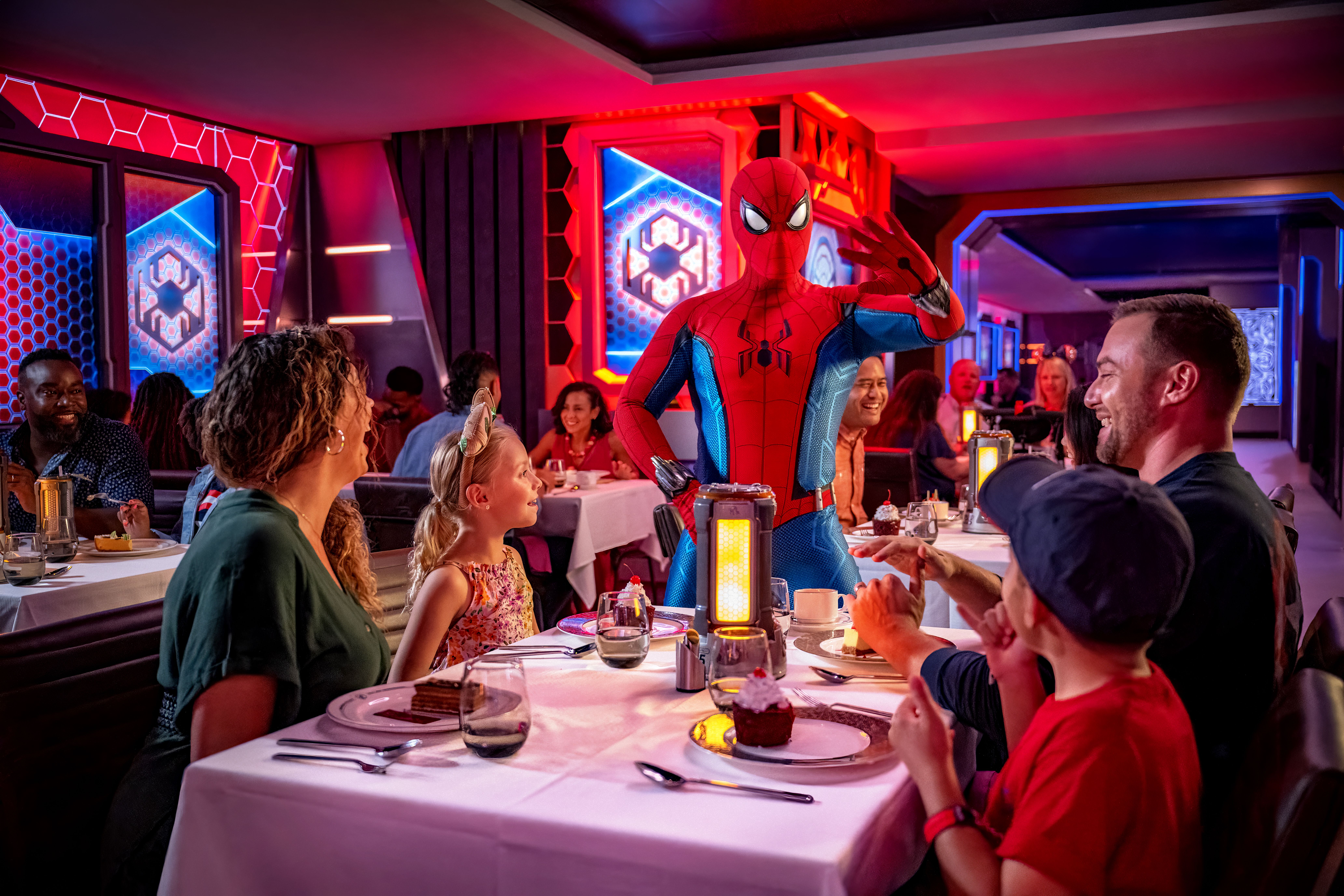 Family dining at Disney Cruise Line's Worlds of Marvel restaurant, interacting with Spider-Man in a futuristic, superhero-themed setting.