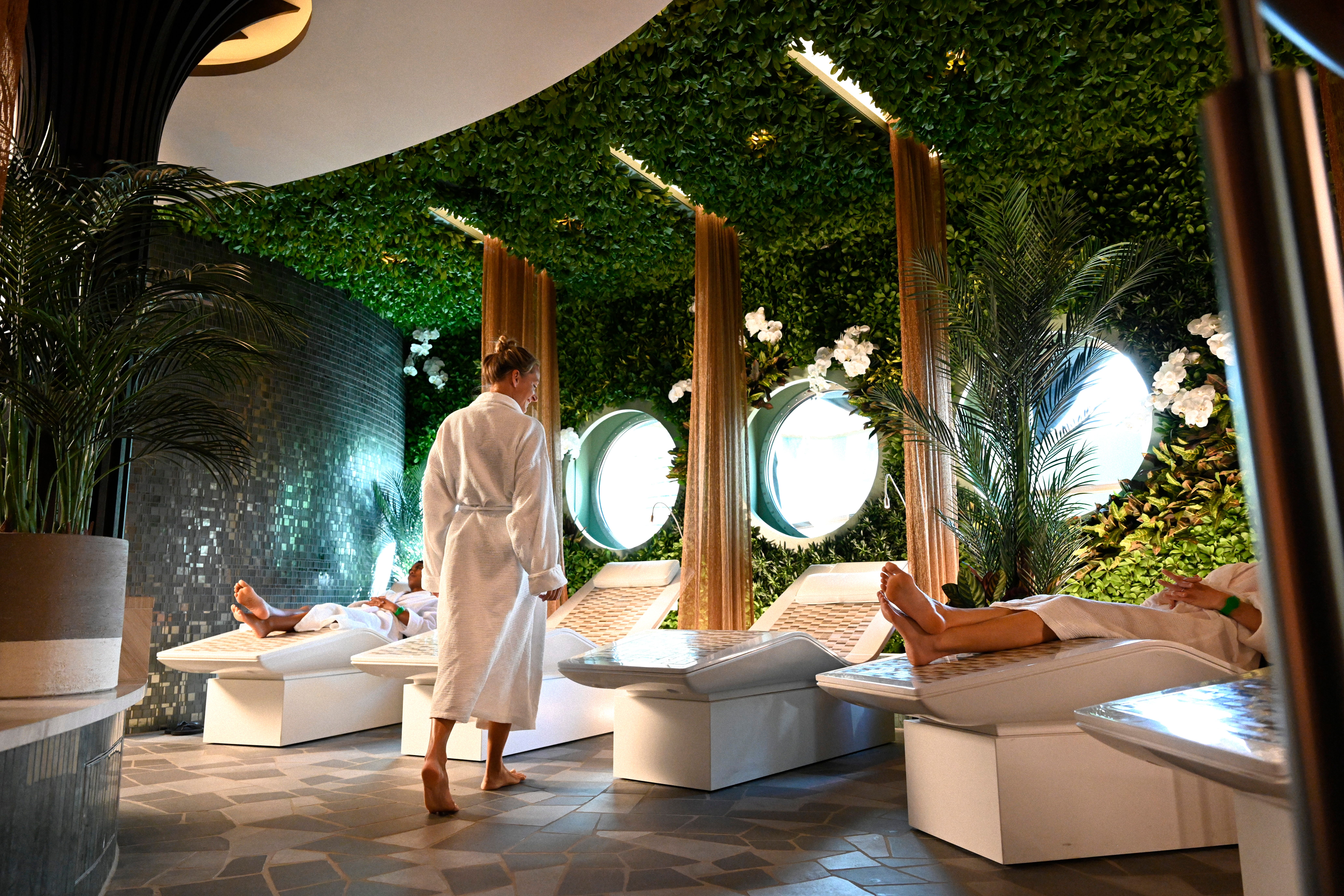 Guests relaxing in a serene, nature-inspired spa aboard Disney Cruise Line, featuring lounge chairs, lush greenery, and soft lighting for a peaceful atmosphere.