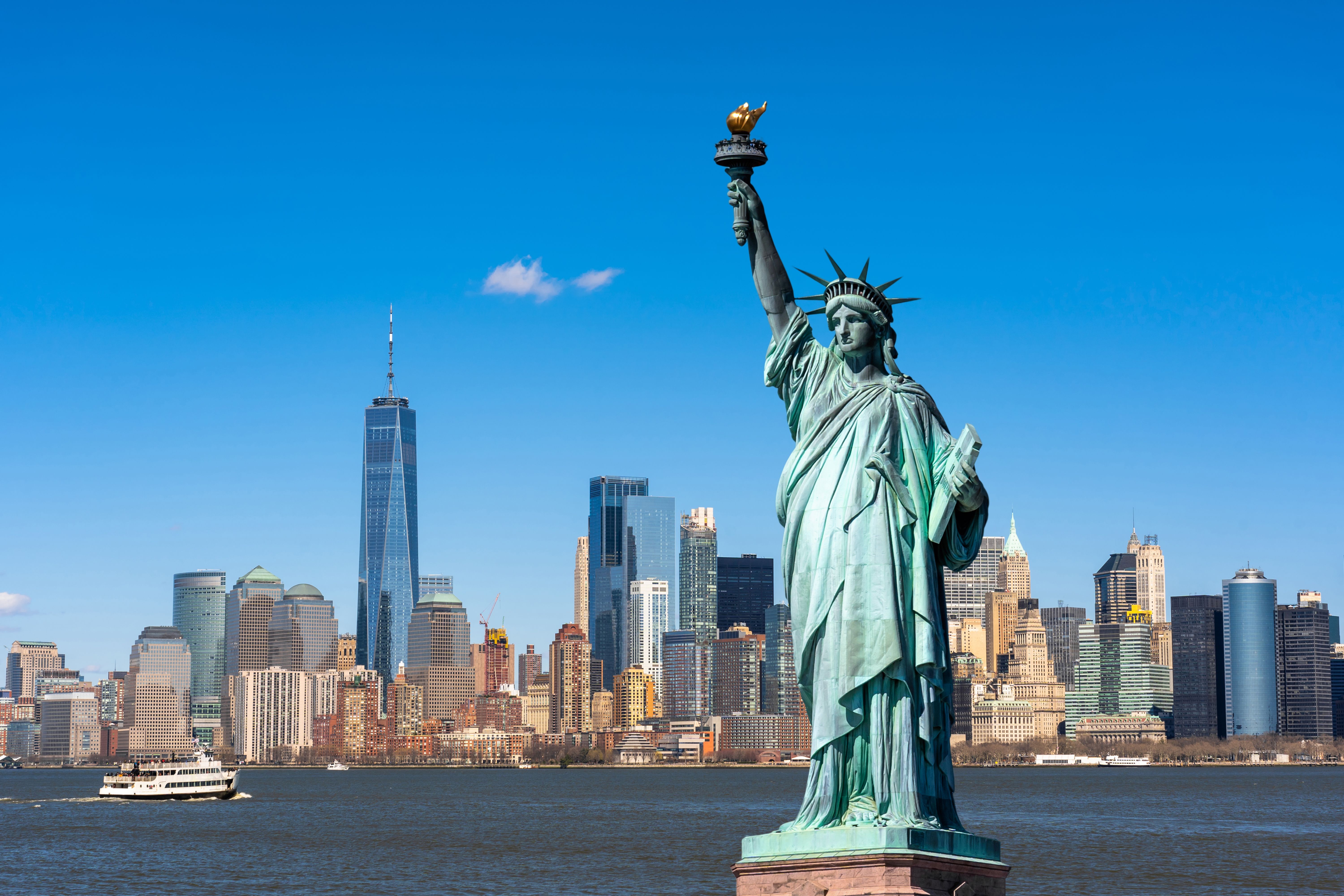 Iconic Statue of Liberty with New York City skyline and One World Trade Center in the background on a clear, sunny day.