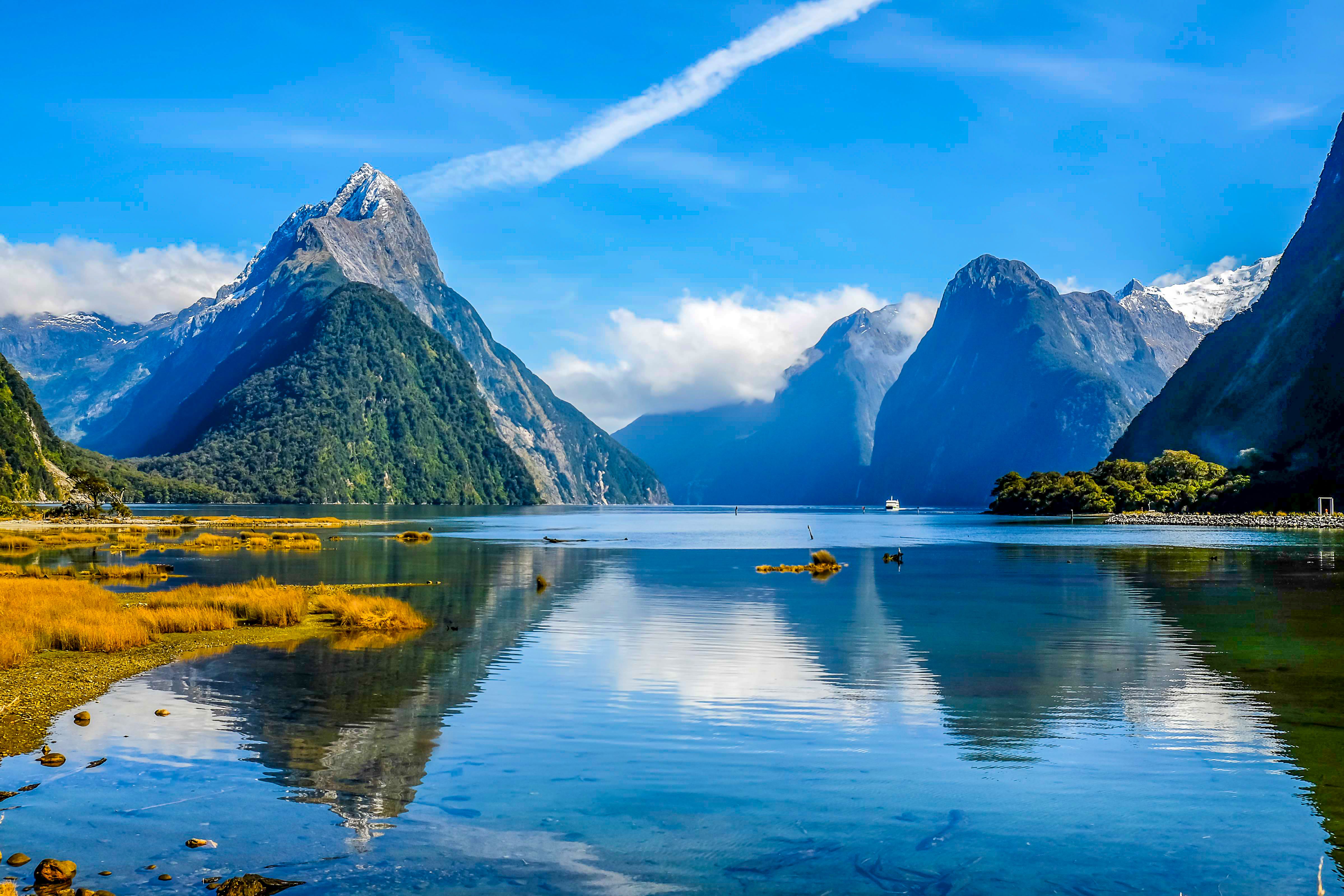 Iconic Milford Sound in New Zealand's Fiordland, featuring Mitre Peak, calm reflective waters, rugged mountains, and lush greenery under a vibrant blue sky.