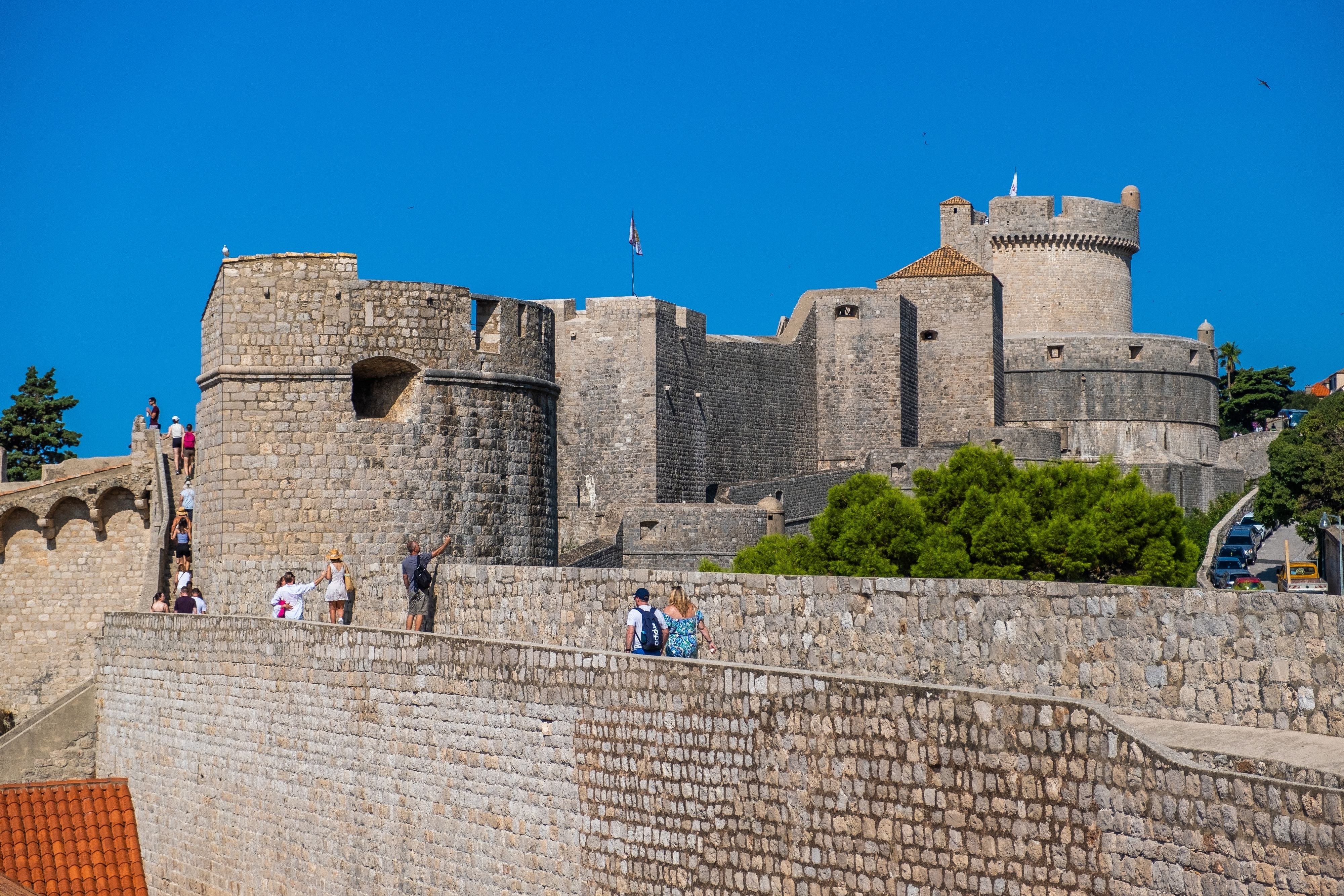 Tourists exploring the ancient stone walls and towers of Dubrovnik's Old Town, Croatia, under a clear blue sky.