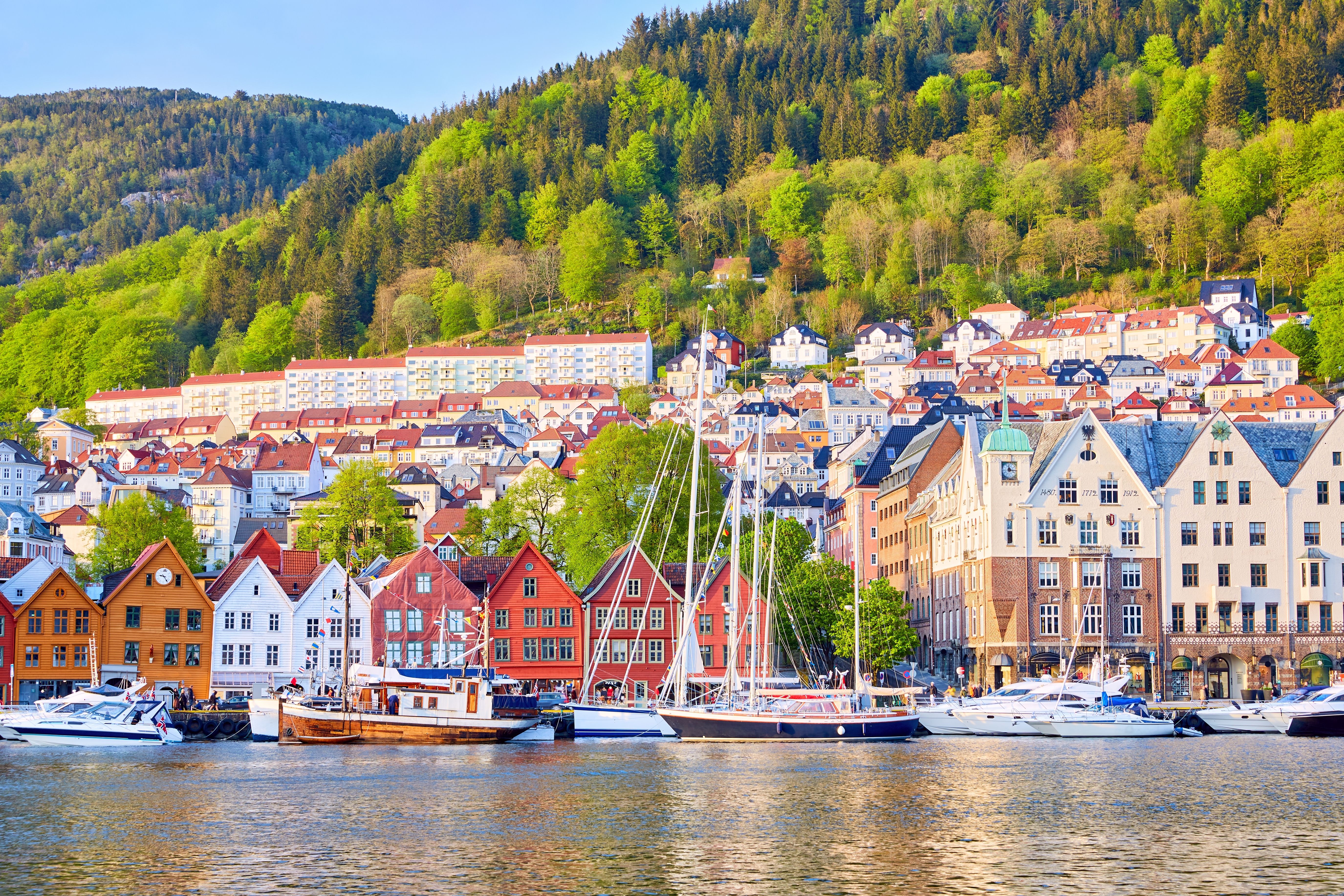 Scenic waterfront of Bryggen in Bergen, Norway, featuring colorful wooden buildings, boats, and a lush green hillside in the background.