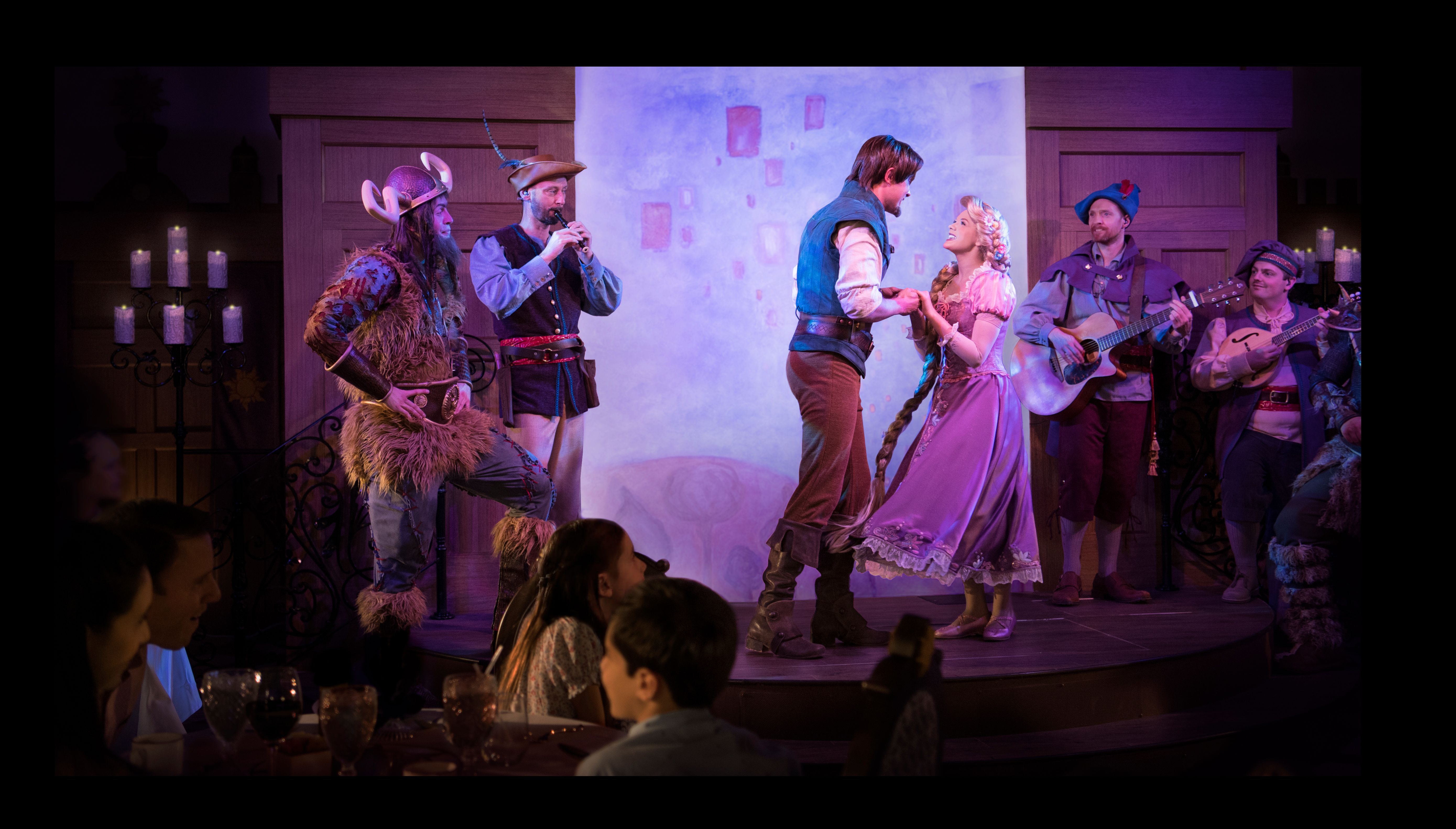 Rapunzel and Flynn Rider dancing on stage during the live dinner show at Rapunzel's Royal Table on the Disney Magic cruise ship, surrounded by performers and festive lantern-themed decor, with diners enjoying the immersive Tangled-themed experience.