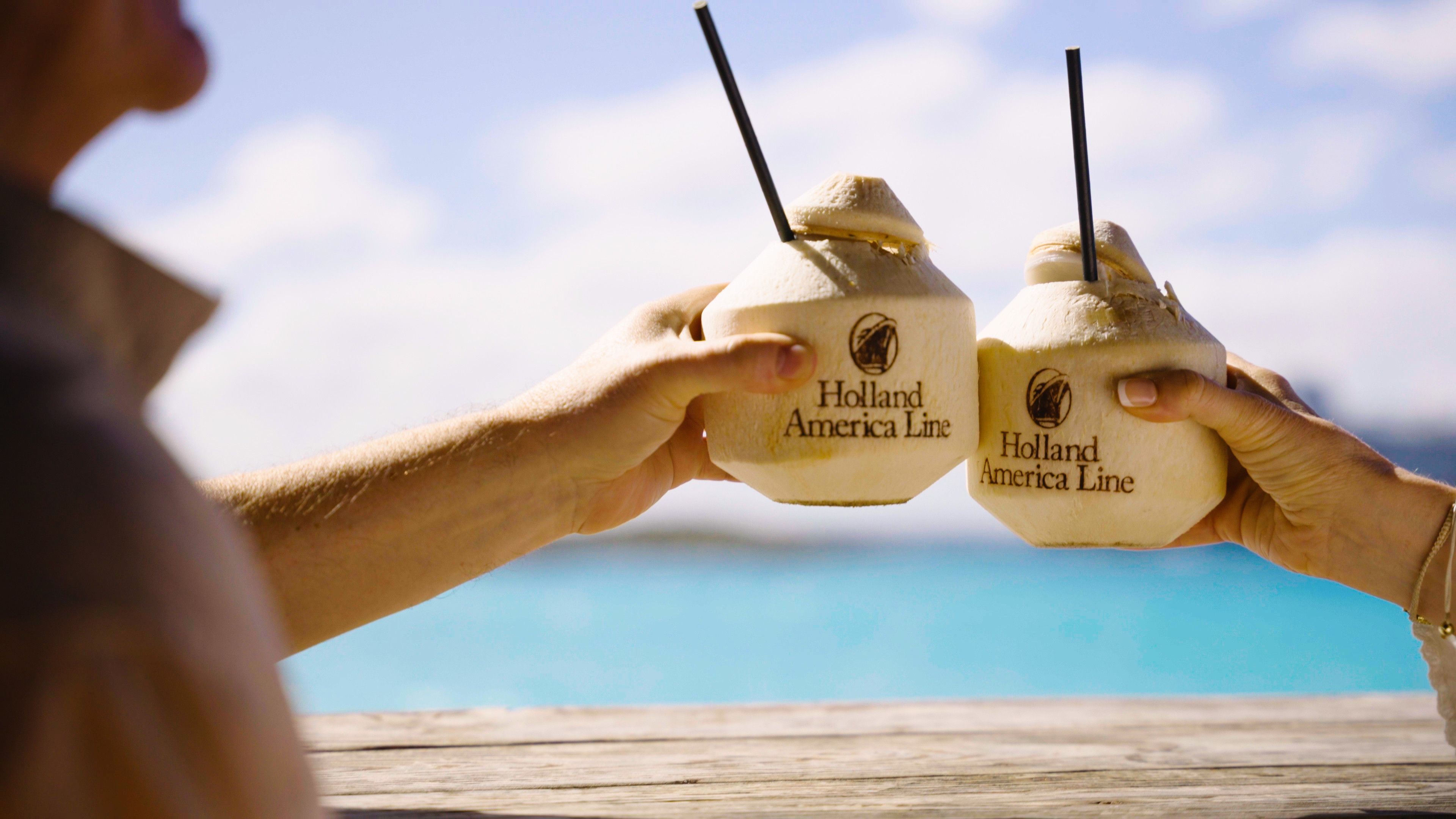 Two hands clinking coconuts with Holland America Line logos, set against a tropical turquoise sea and sky, symbolizing a celebratory cruise moment.