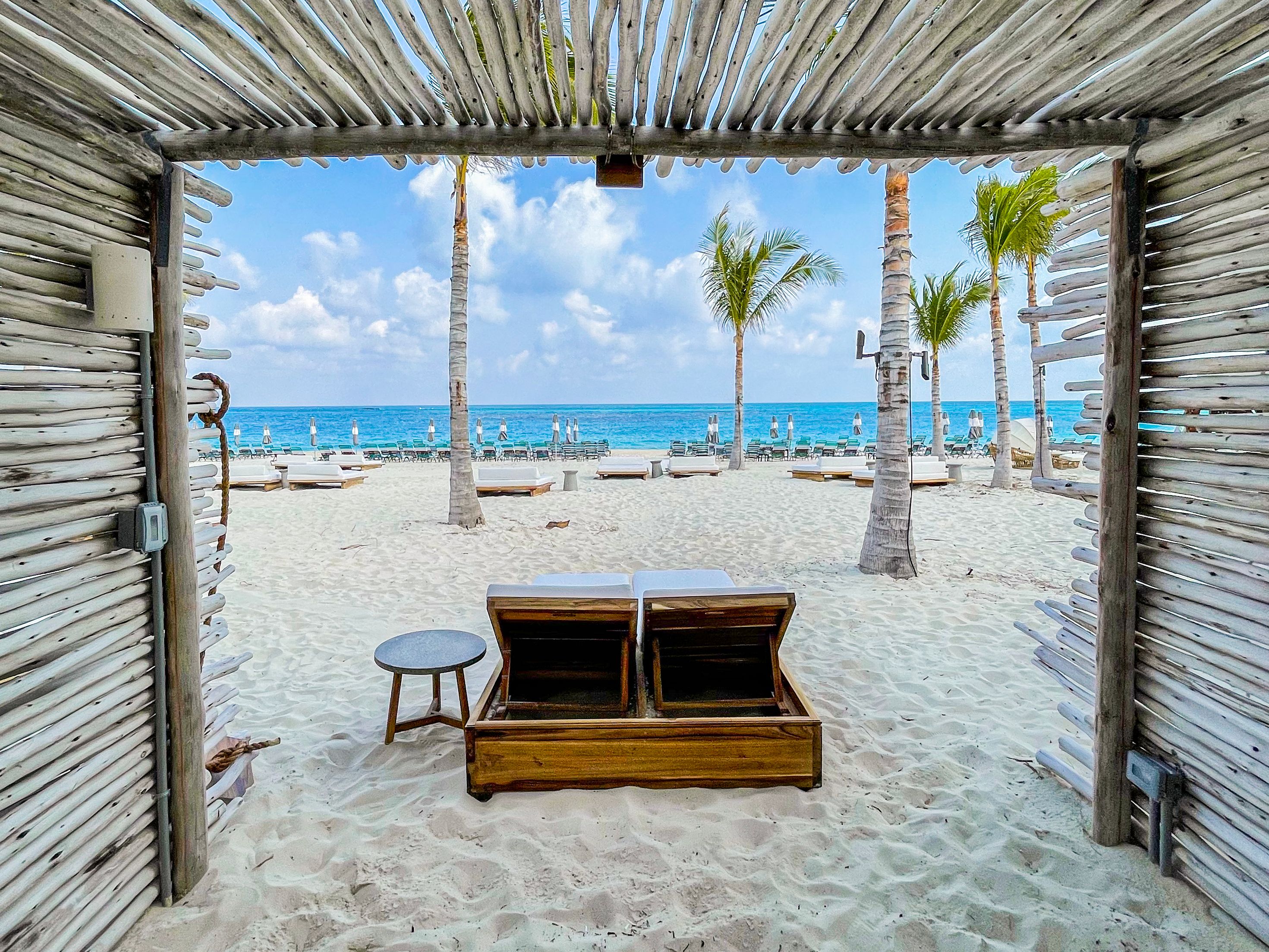 View from a wooden cabana at Bimini Beach Club, featuring two lounge chairs, a table, palm trees, and a pristine beach leading to the turquoise ocean.