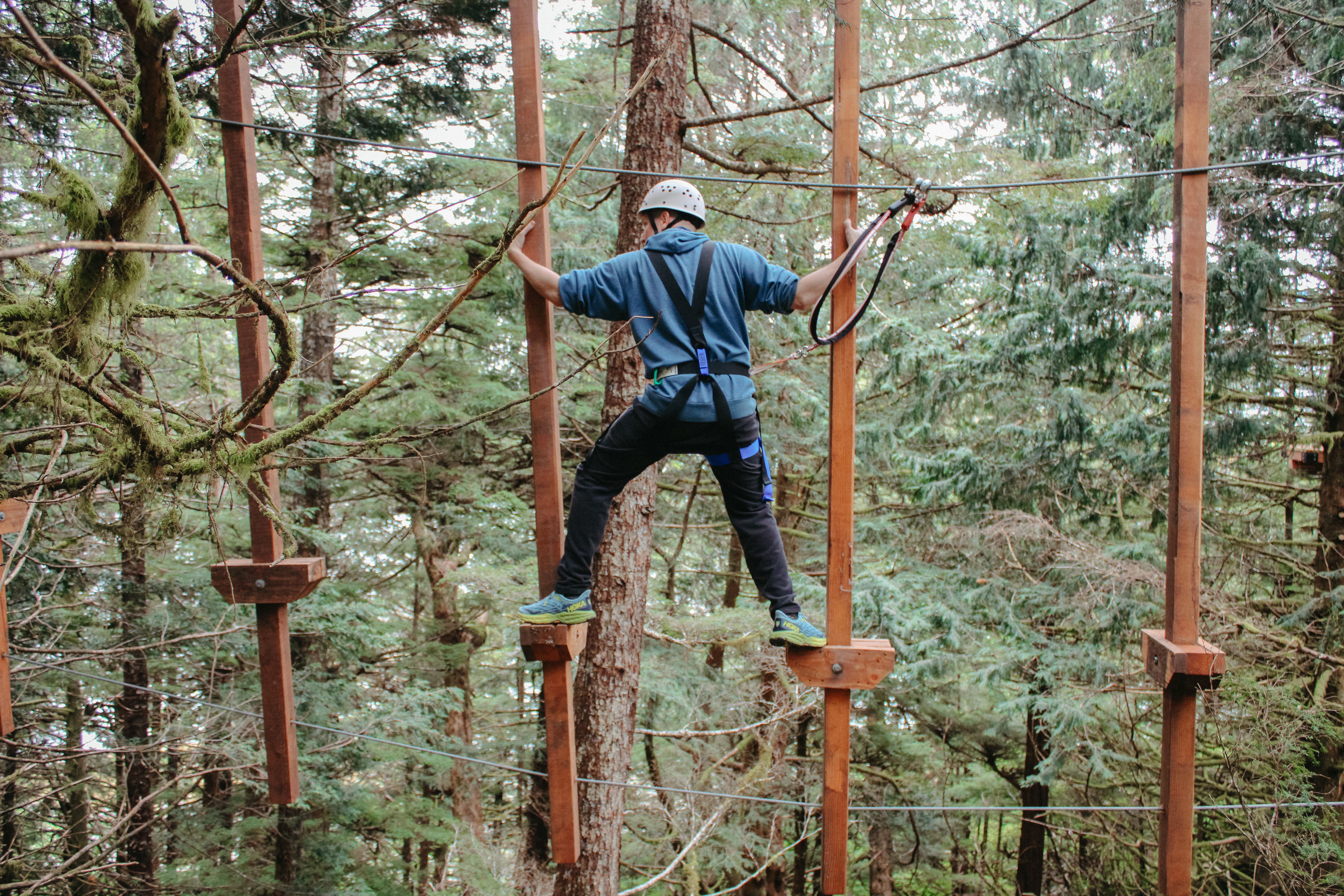 Person navigating a treetop aerial ropes course in a lush forest, wearing a harness and helmet for safety, balancing on wooden platforms connected by cables.