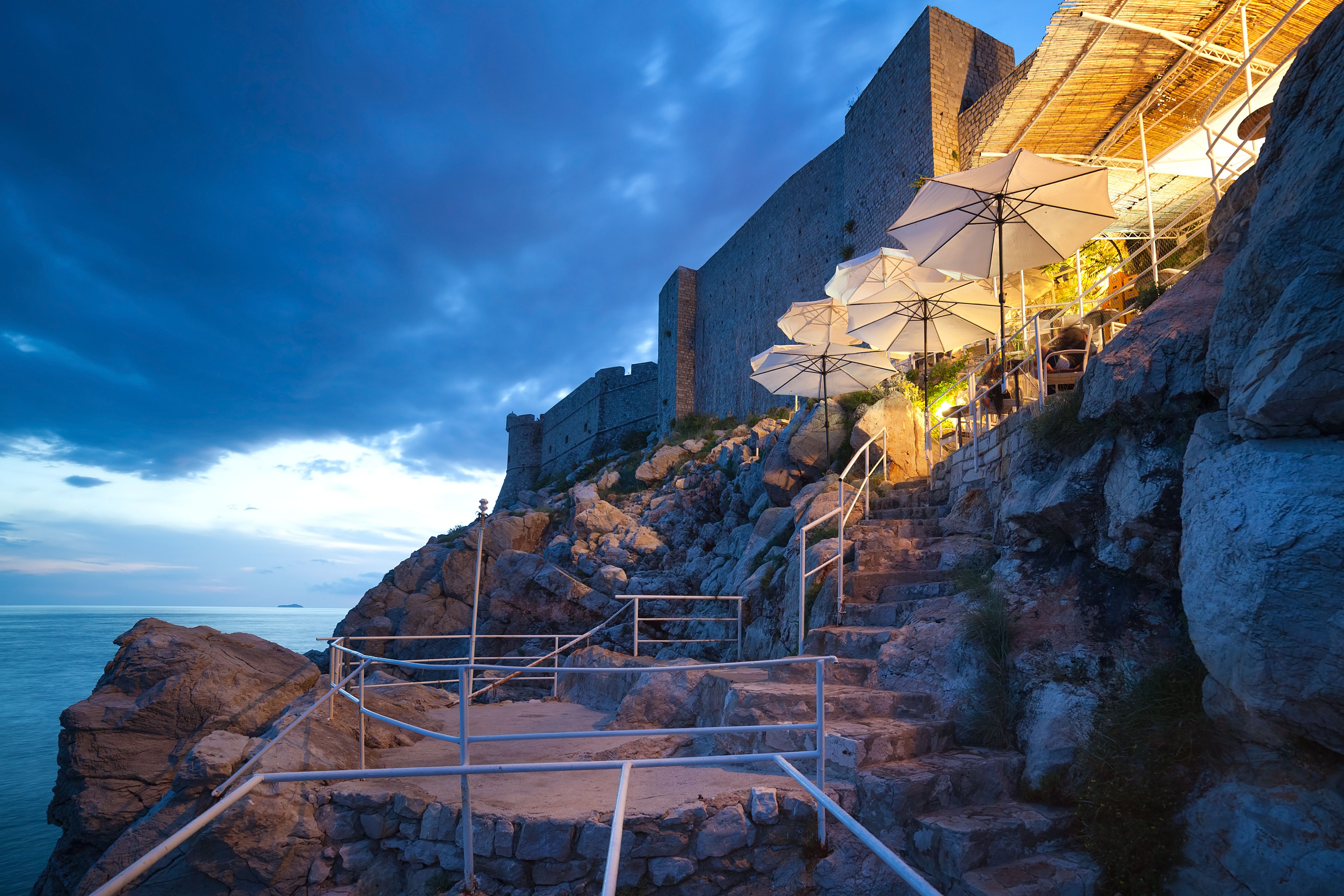 Cliffside bar in Dubrovnik, Croatia, with outdoor seating and umbrellas overlooking the Adriatic Sea at dusk, offering a scenic and romantic atmosphere.