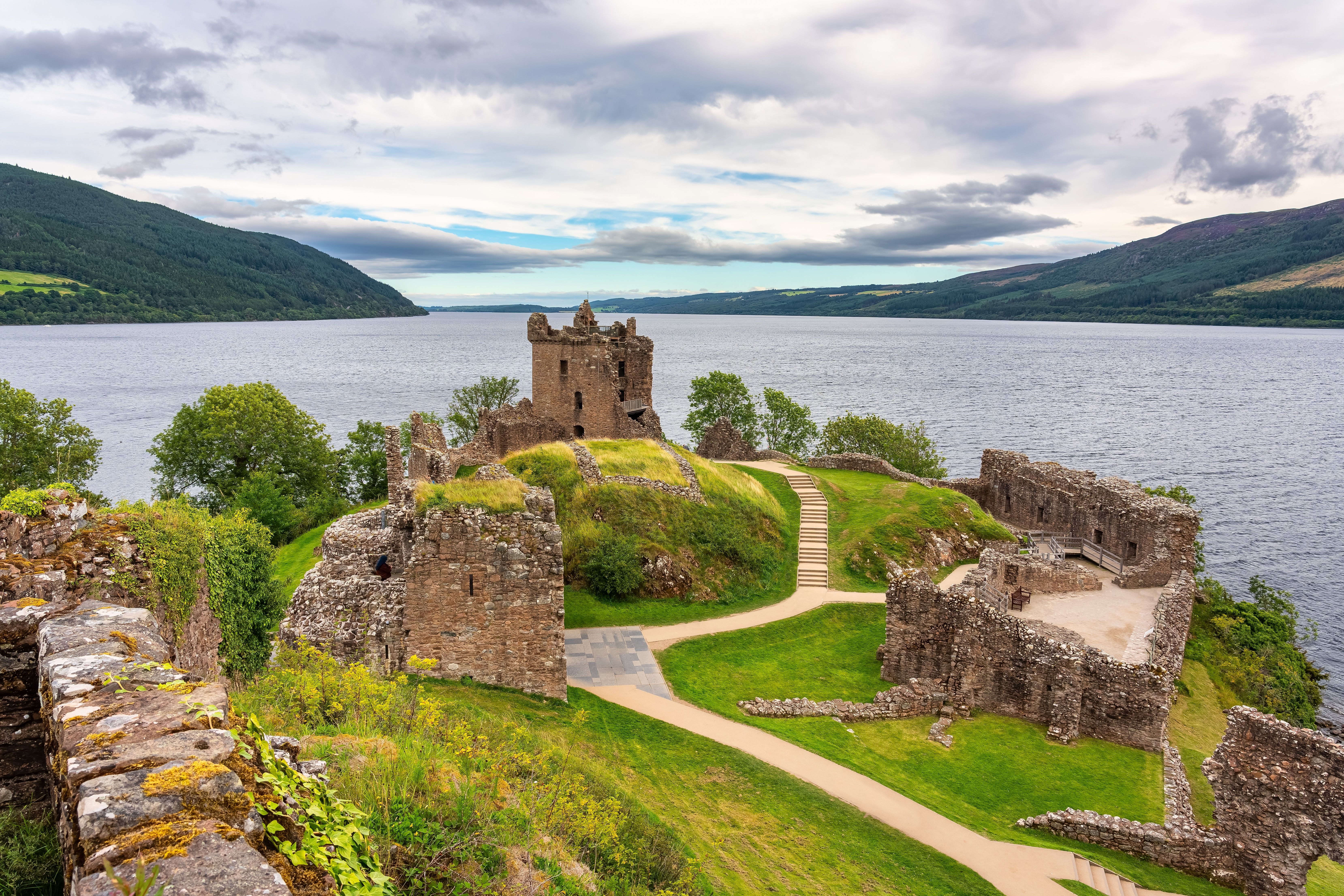 Urquhart Castle ruins overlooking Loch Ness in the Scottish Highlands, surrounded by lush greenery and rolling hills under a cloudy sky.