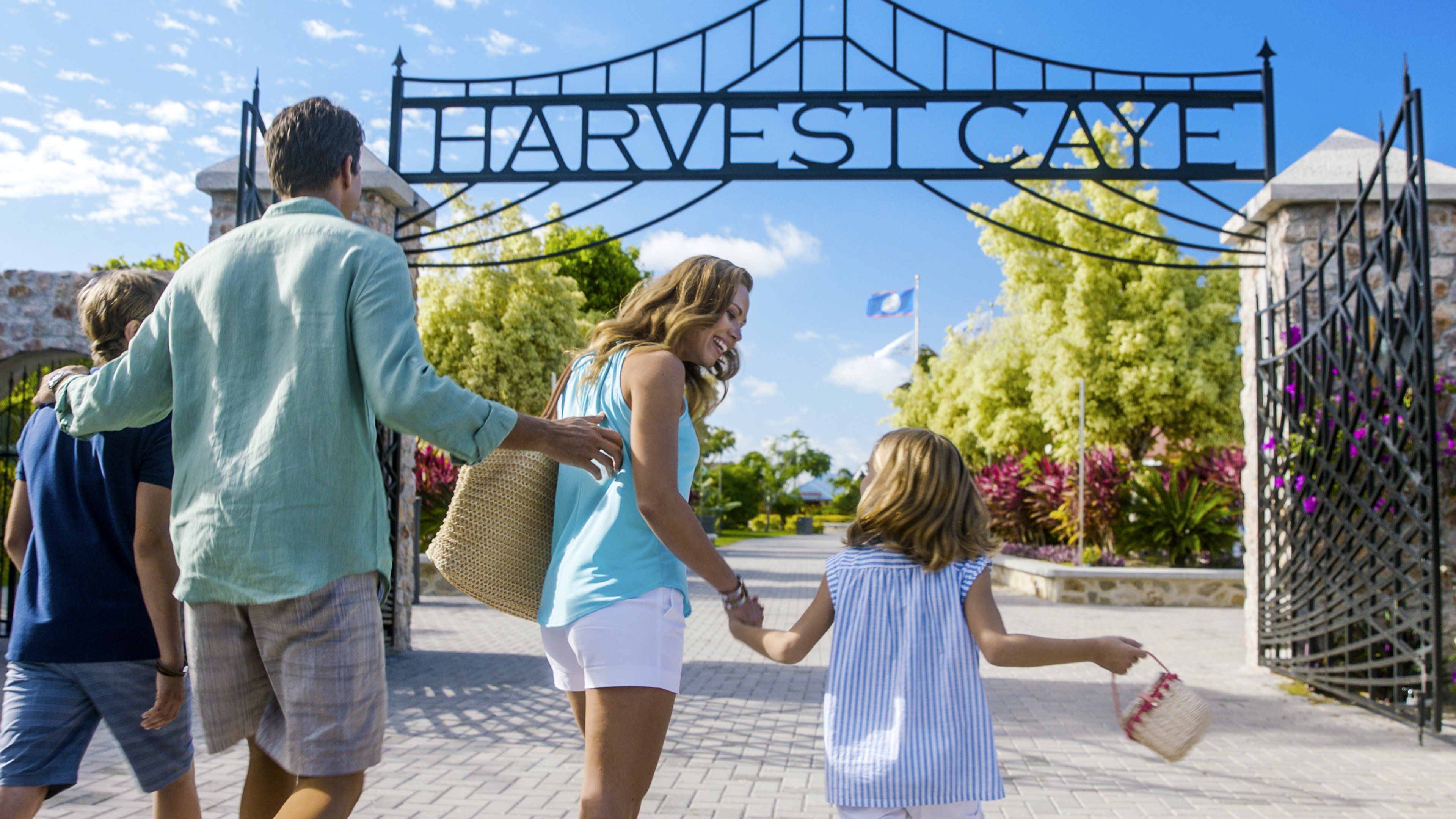Family entering Harvest Caye, Belize through an ornate gate with tropical plants and blue skies.