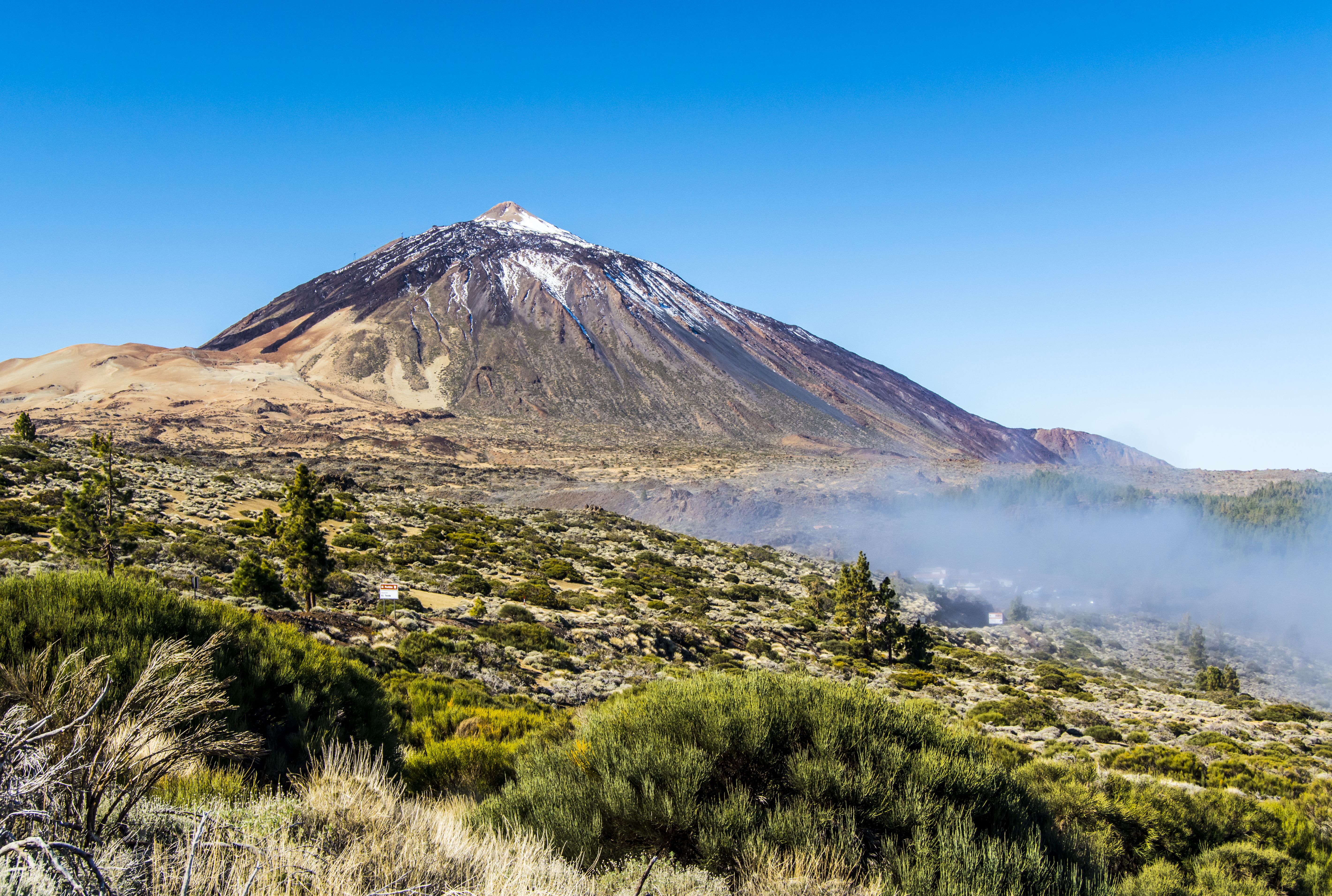 Mount Teide in Tenerife, Canary Islands, with rugged slopes, sparse vegetation, and a snow-capped peak under a clear blue sky.