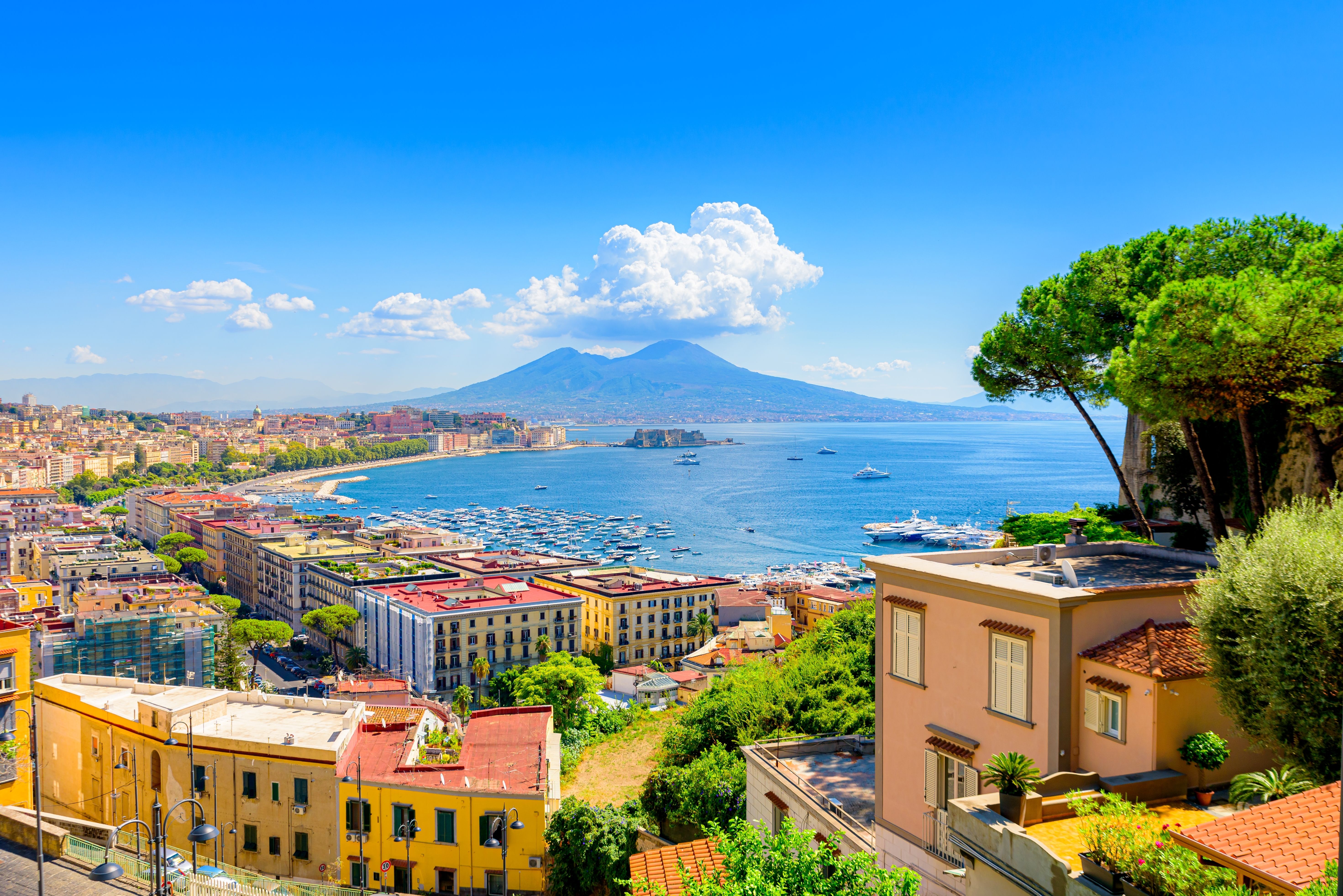 Scenic view of Naples, Italy, with colorful buildings, a busy harbor, and Mount Vesuvius in the background under a clear blue sky.