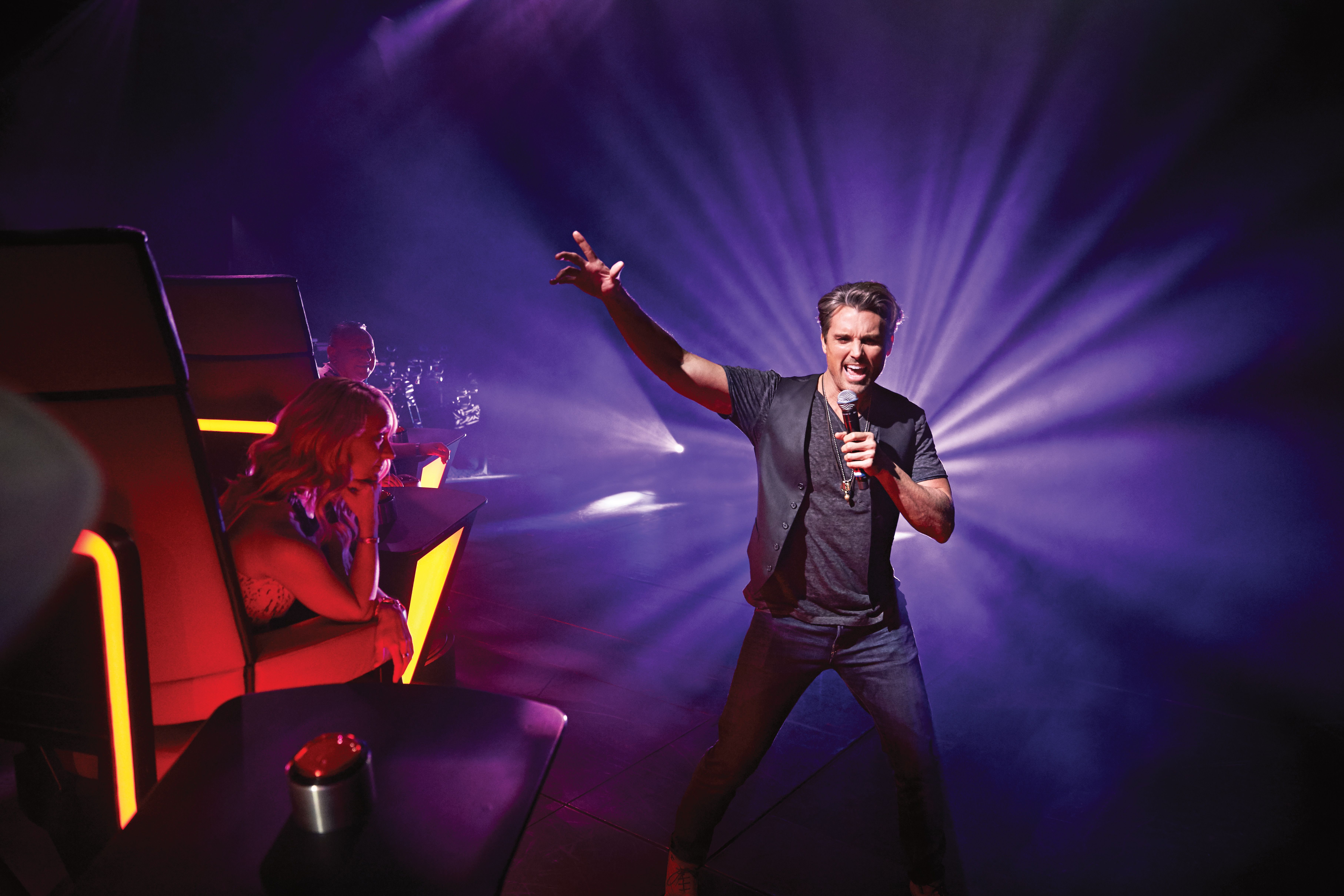 The Voice of the Ocean live show on Princess Cruises featuring a contestant performing on stage with illuminated judge chairs in dramatic lighting.