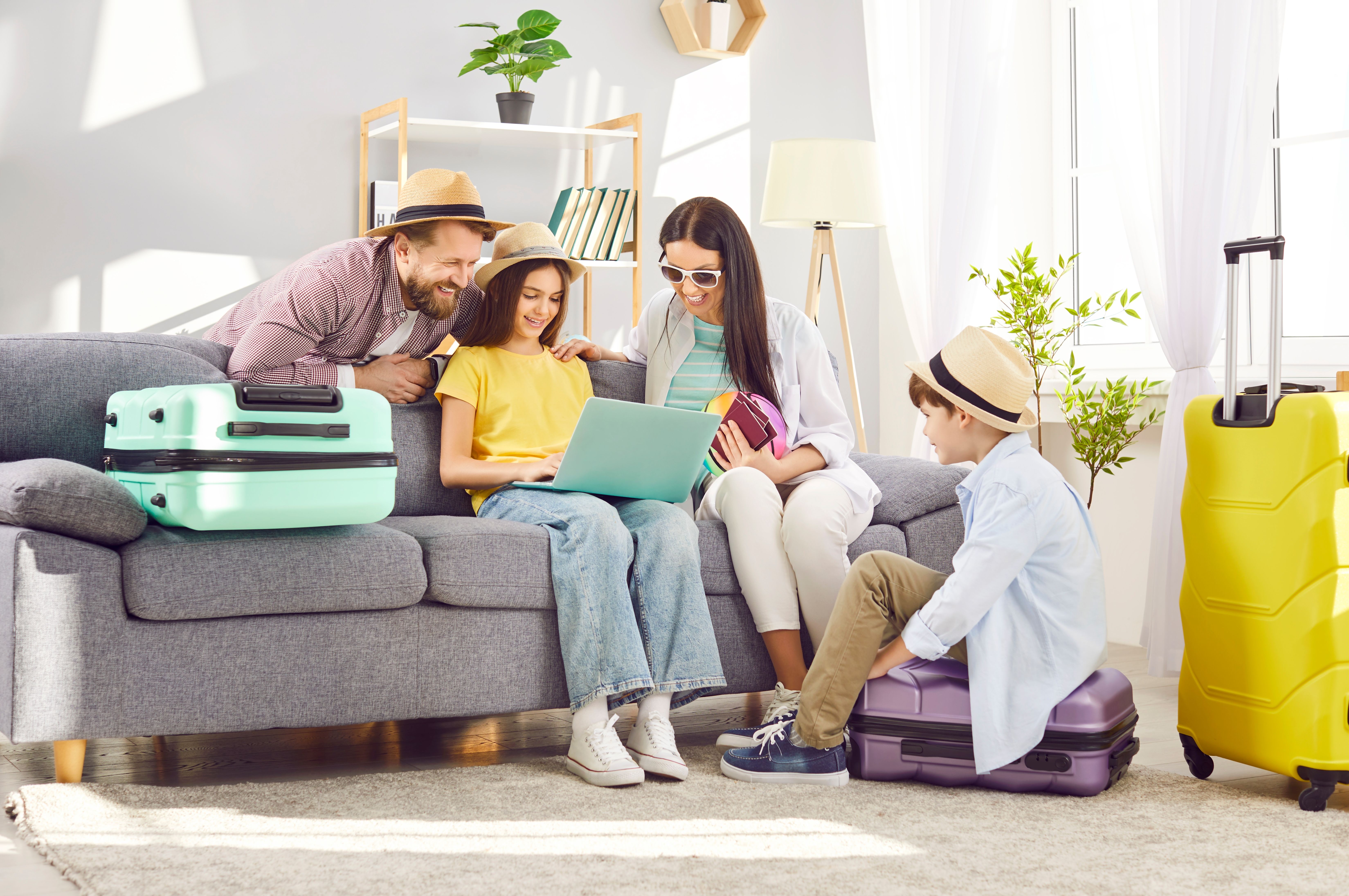 Excited family with luggage planning a cruise vacation at home, using laptop while packing in bright, sunlit living room.
