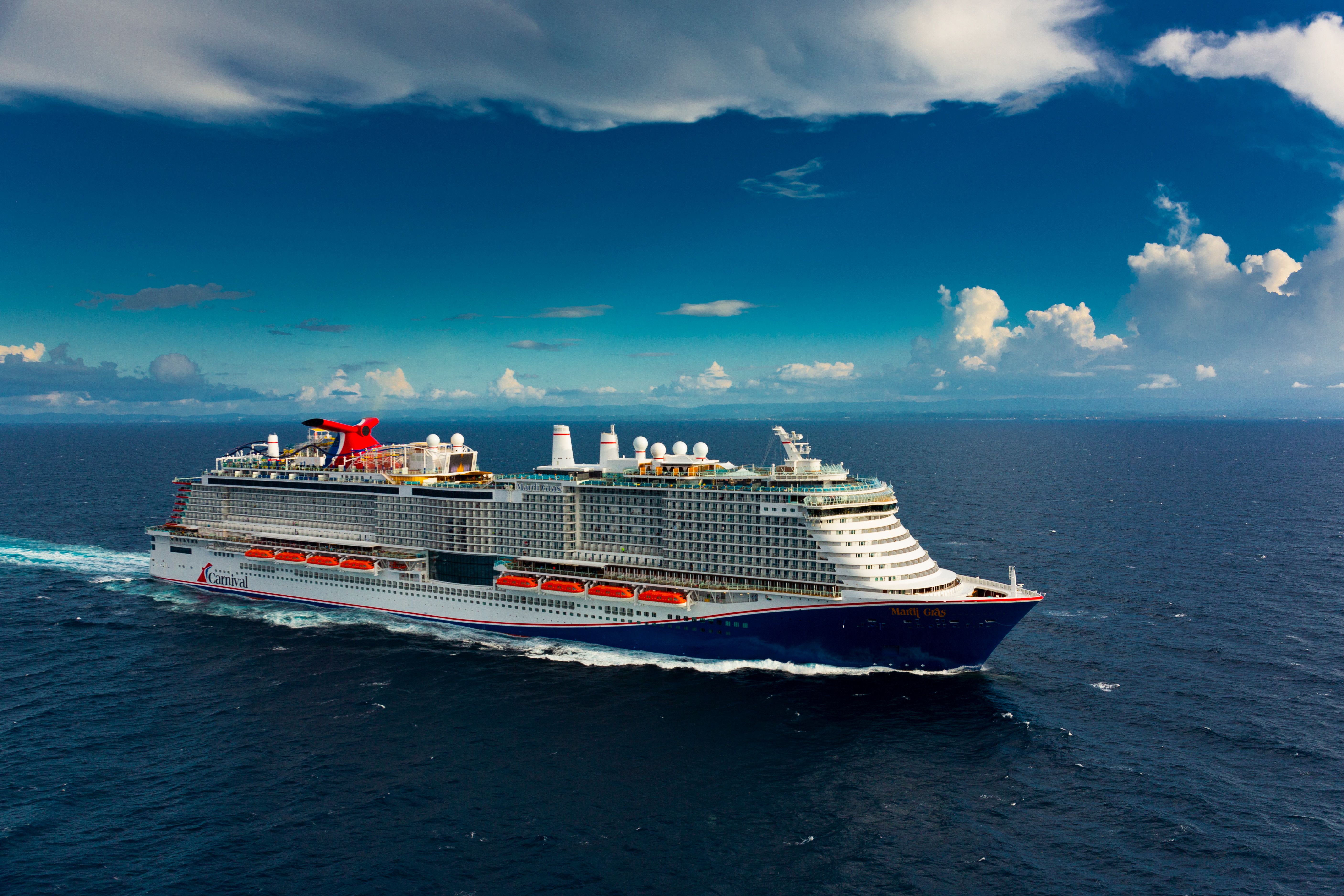 Carnival Mardi Gras sailing through open ocean under blue sky, showcasing red funnel, upper decks, and balcony staterooms.